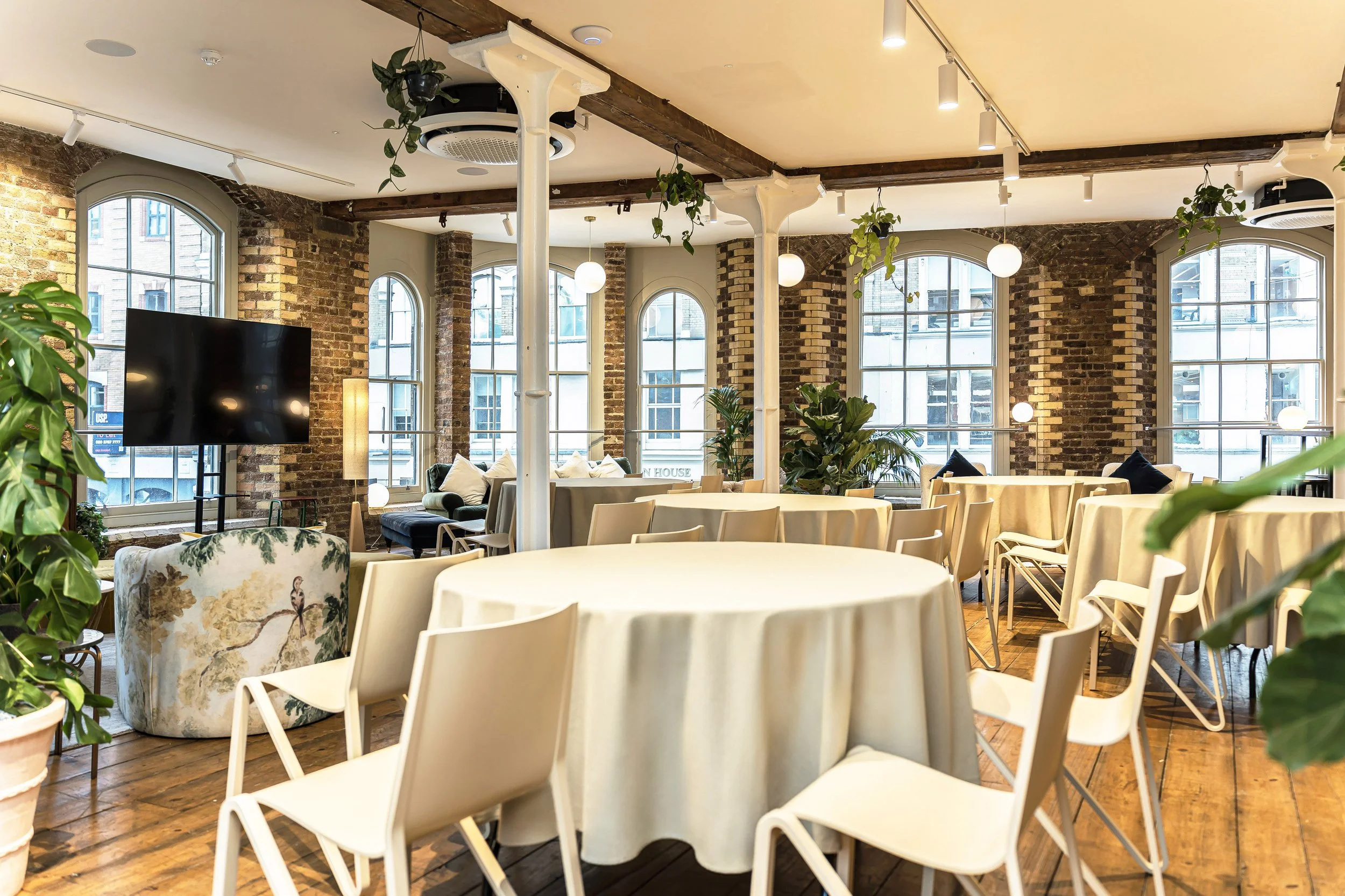 Bright, spacious room with large arched windows, exposed brick walls, and a wooden ceiling. The room features round tables with cream-colored tablecloths, surrounded by white chairs. There are various plants, a large flat-screen TV, and pendant and t
