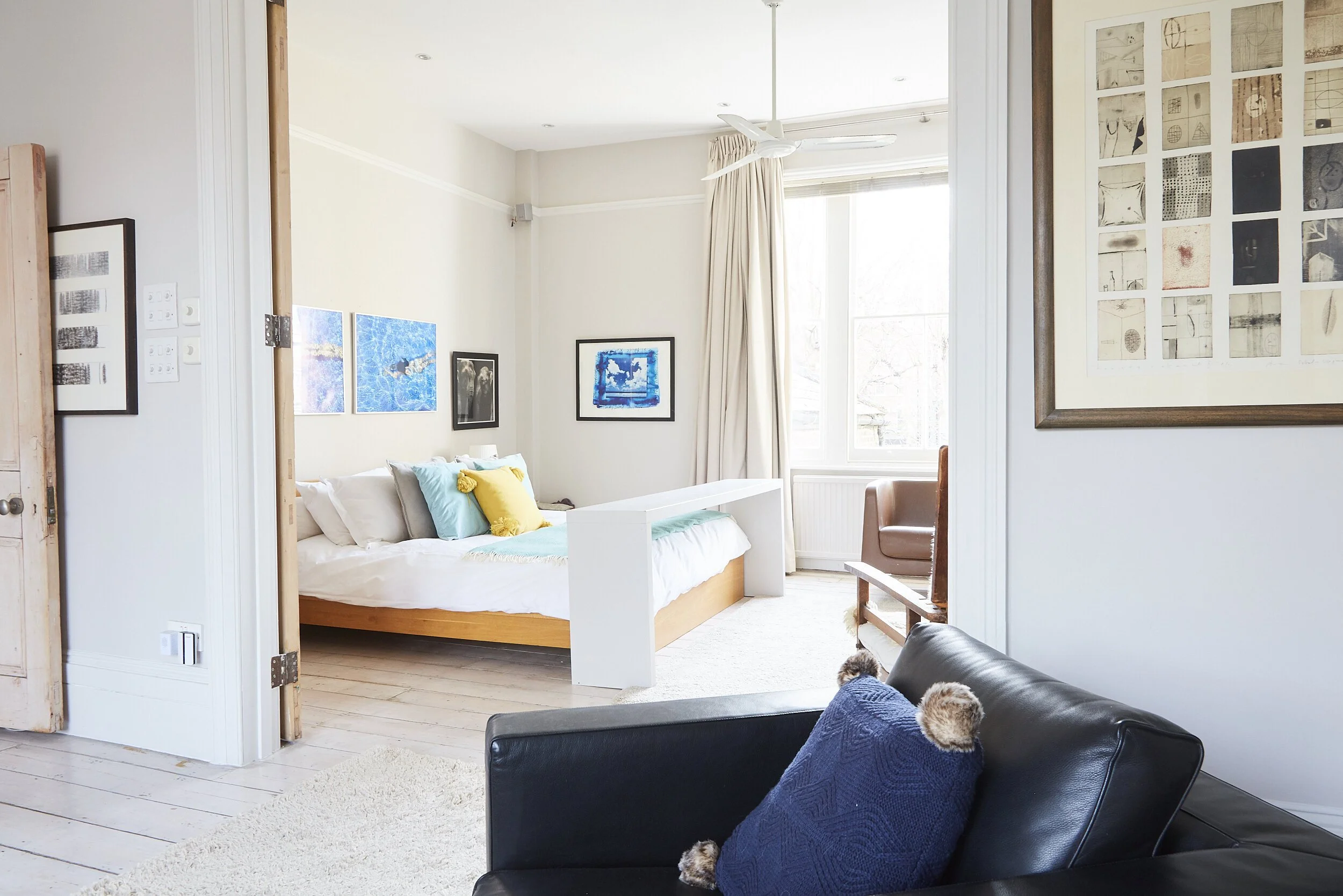 A bright bedroom with a bed decorated with colorful pillows, artwork on the walls, a window with curtains, and a leather chair with a pillow in the foreground.