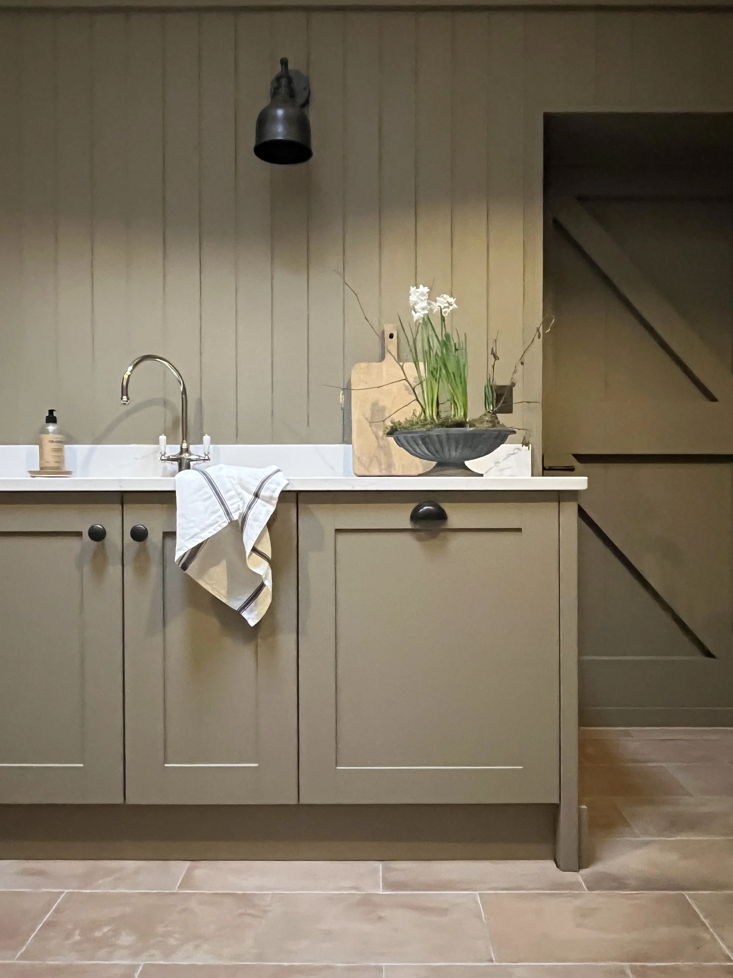 Kitchen sink area with beige cabinets, white countertop, chrome faucet, soap dispenser, white dish towel hanging on the cabinet handle, potted plant with white flowers on the counter, cutting board behind the plant, black wall-mounted light fixture, 