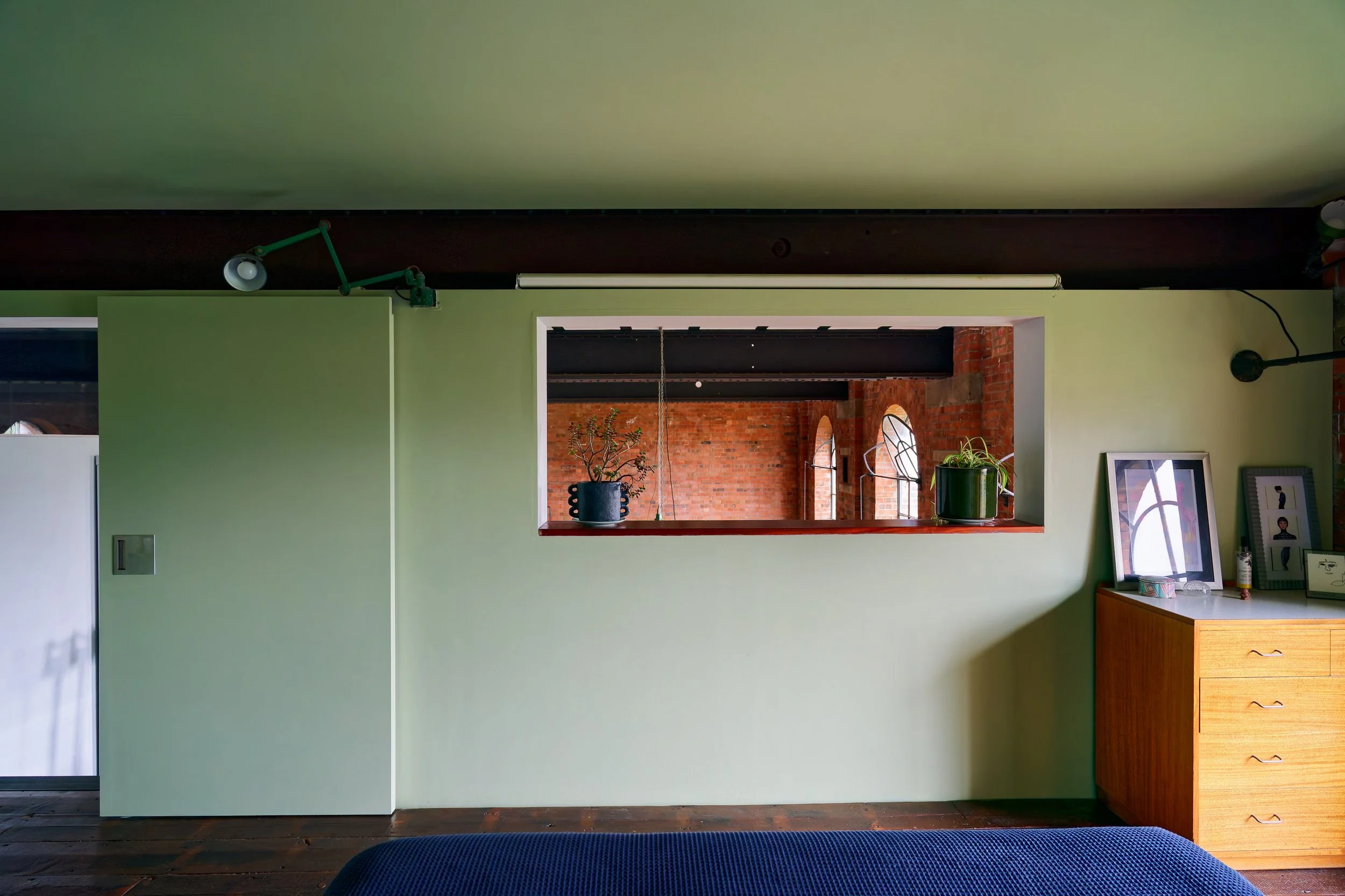A view of a room with green walls, a wooden dresser with framed photos and a bottle, potted plants on a ledge, and exposed brick walls visible through an open window.