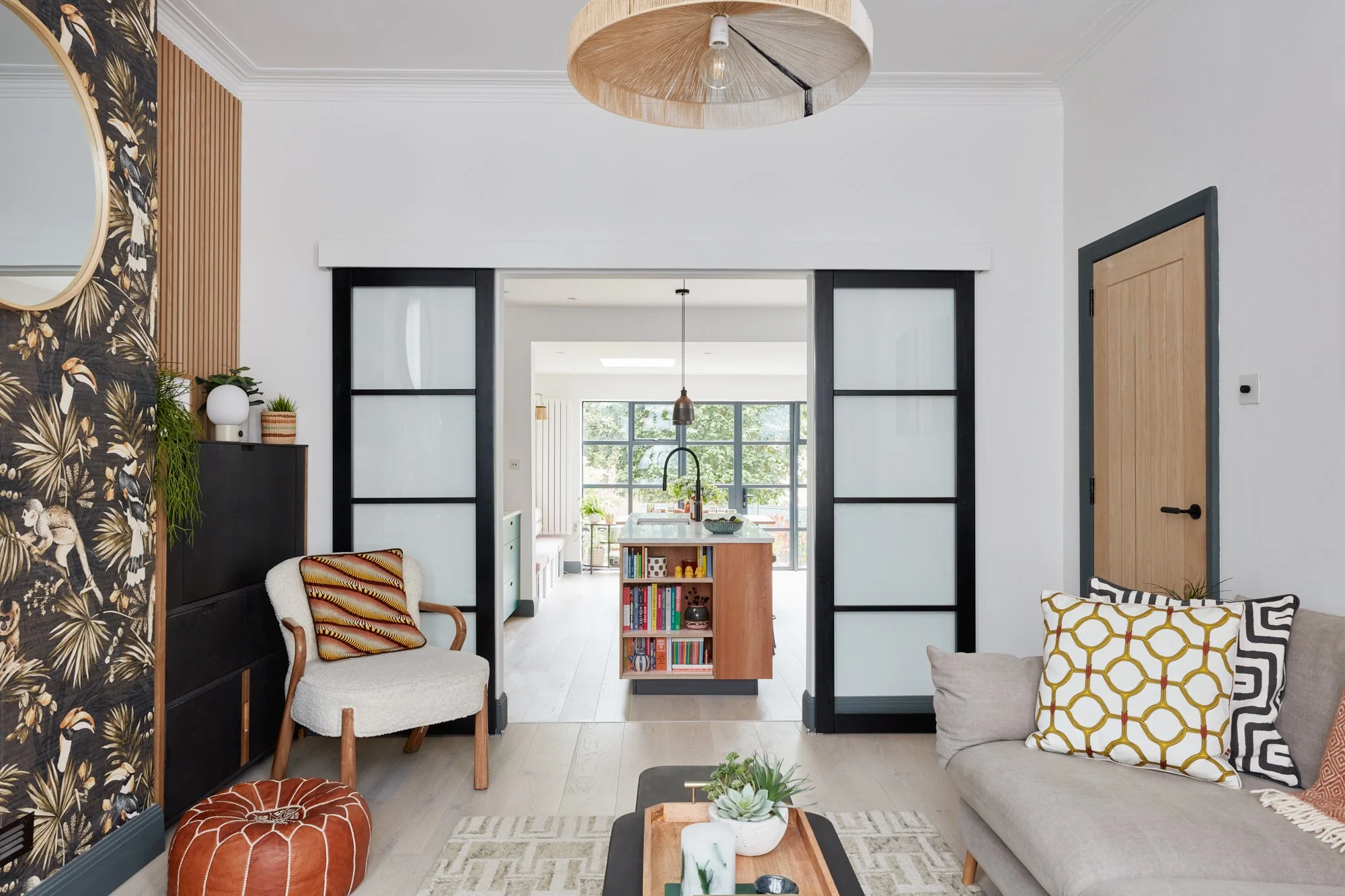 Living room with patterned wallpaper, white and beige furniture, decorative pillows, and a view into a bright kitchen with large windows.