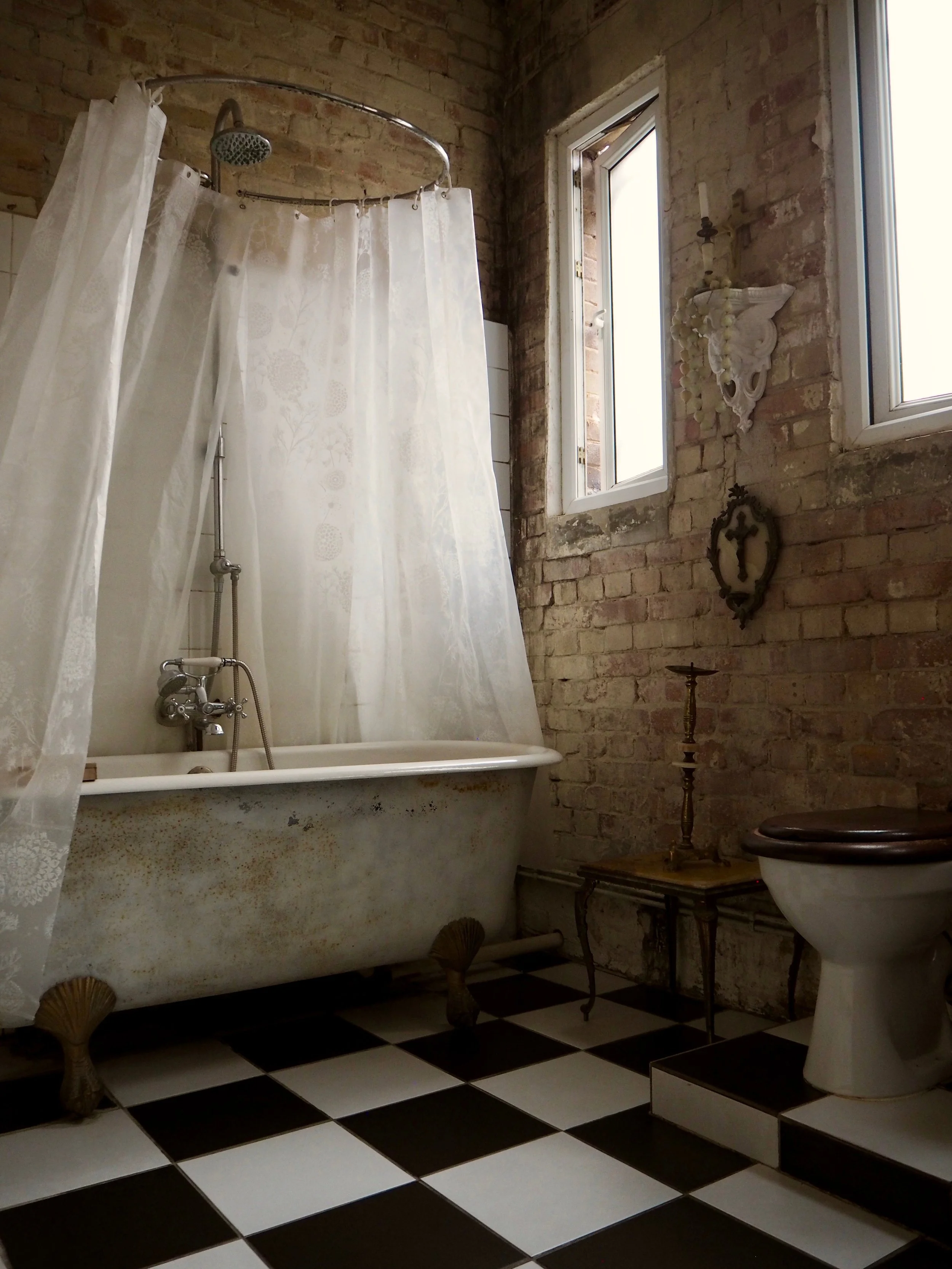 Bathroom with vintage clawfoot tub partially enclosed with a sheer, floral shower curtain, black and white checkered floor, exposed brick walls, two small windows, and antique decor.