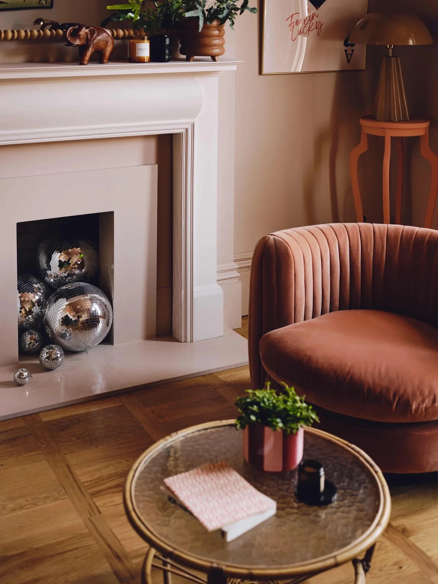 A living room scene featuring a white fireplace with shiny disco balls inside, a pink velvet armchair, a small round glass-top side table with a pink potted plant, a pink book, and black glasses, and a framed motivational poster on the wall.