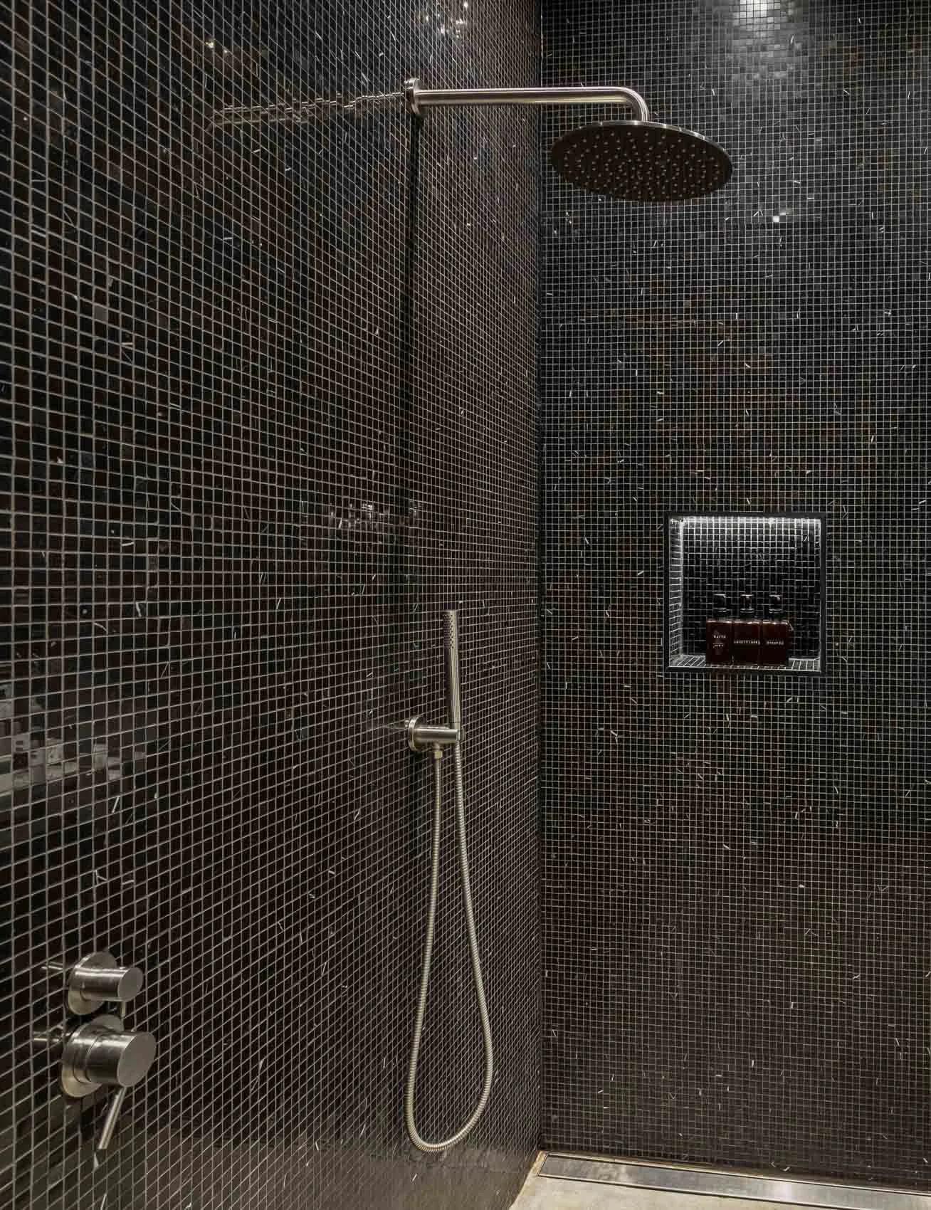 A minimalist black-tiled walk-in shower with a rain showerhead, a handheld shower, and a recessed shelf holding bottles.
