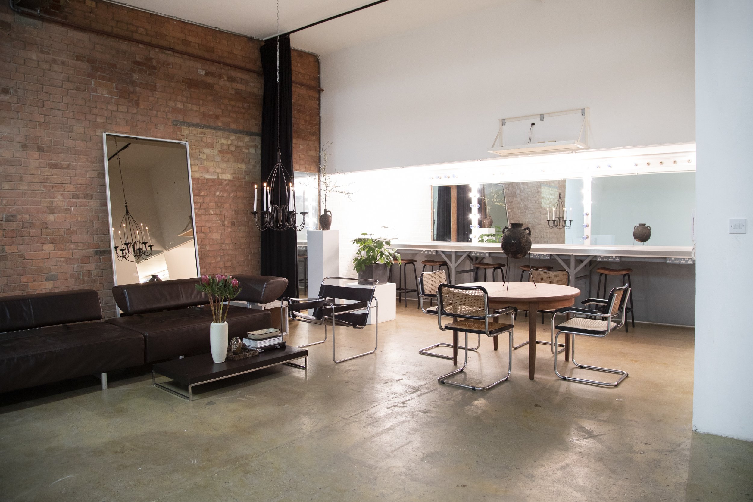 Interior of a modern, minimalist lounge with a brick wall, large mirror, leather sofa, chairs, potted plants, and a vanity mirror surrounded by lights.