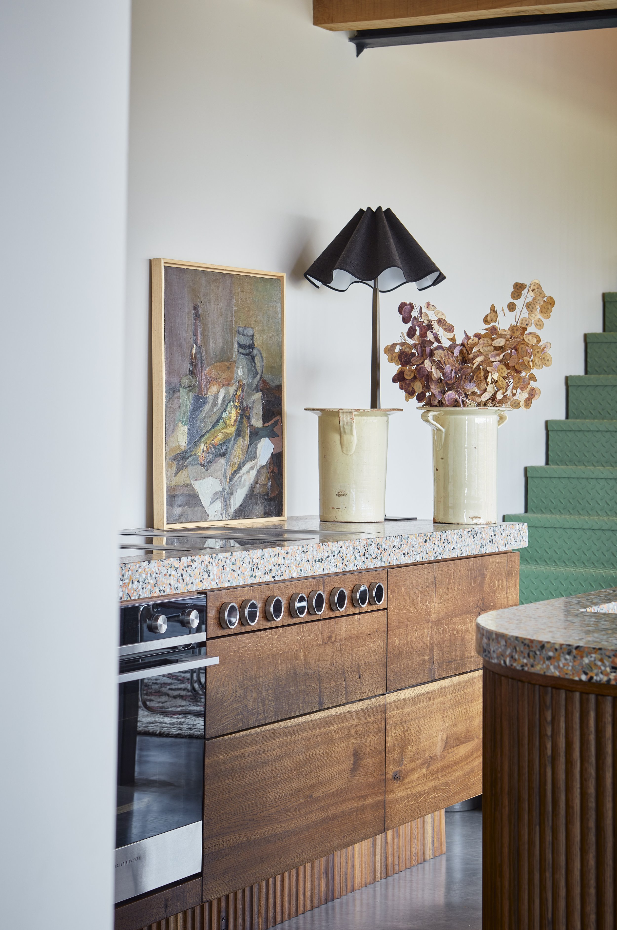 A kitchen with wooden cabinetry, a speckled countertop, a black stove, two cream-colored vases with dried plants, a black-lamped table, a framed abstract painting, and a set of green stairs.