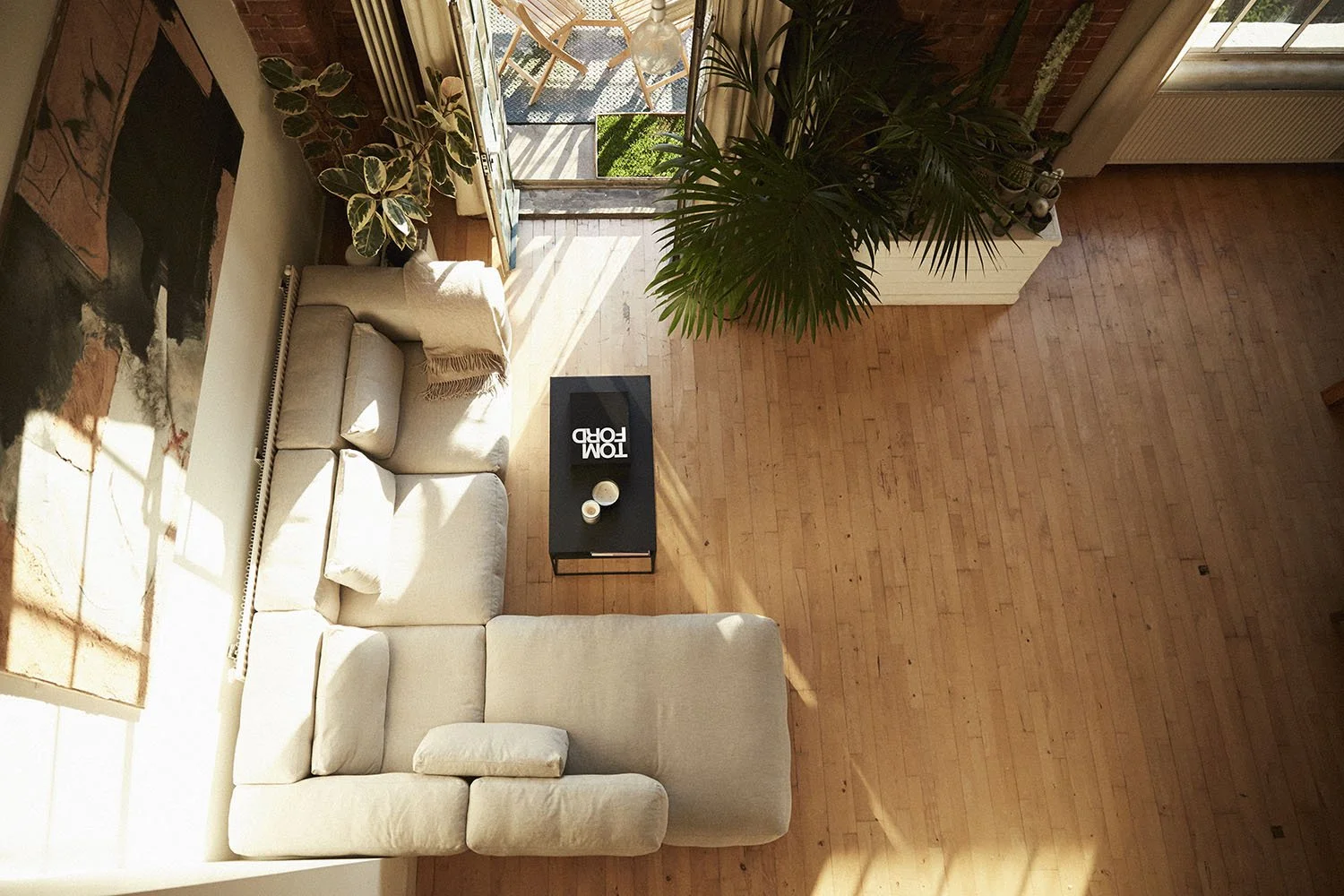 A living room viewed from above with a beige sectional sofa, a black coffee table with decorative items, a large green plant, and an open door leading to a balcony with outdoor chairs and rug, sunlight streaming through.
