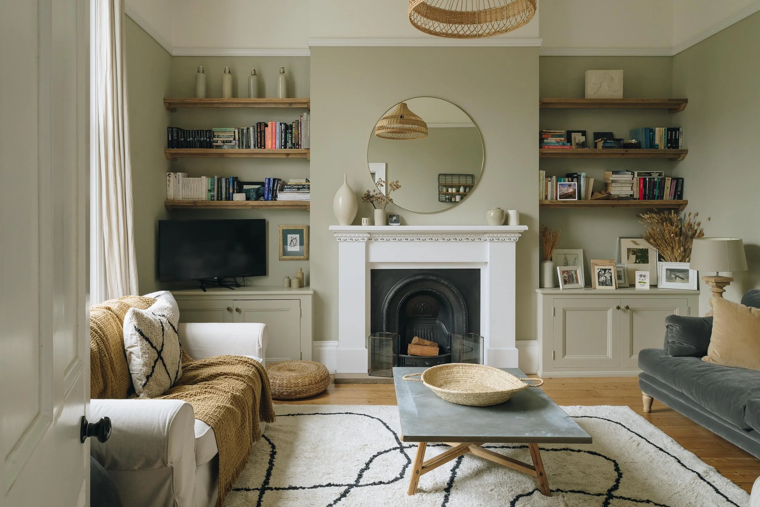 Living room with a fireplace, surrounded by built-in bookshelves, a flat-screen TV, a cream-colored sofa with a blanket, a gray sofa, a coffee table with a round basket, a rug with black lines, decorative items on shelves, and framed photos and dried