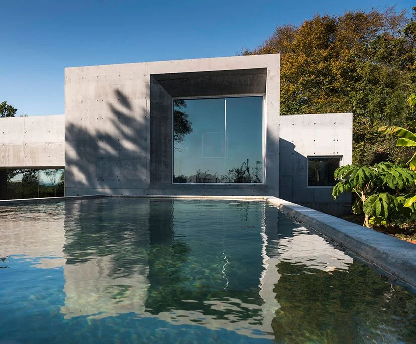 Modern concrete house with large glass windows reflected in a swimming pool, surrounded by trees and shrubs under a clear blue sky.