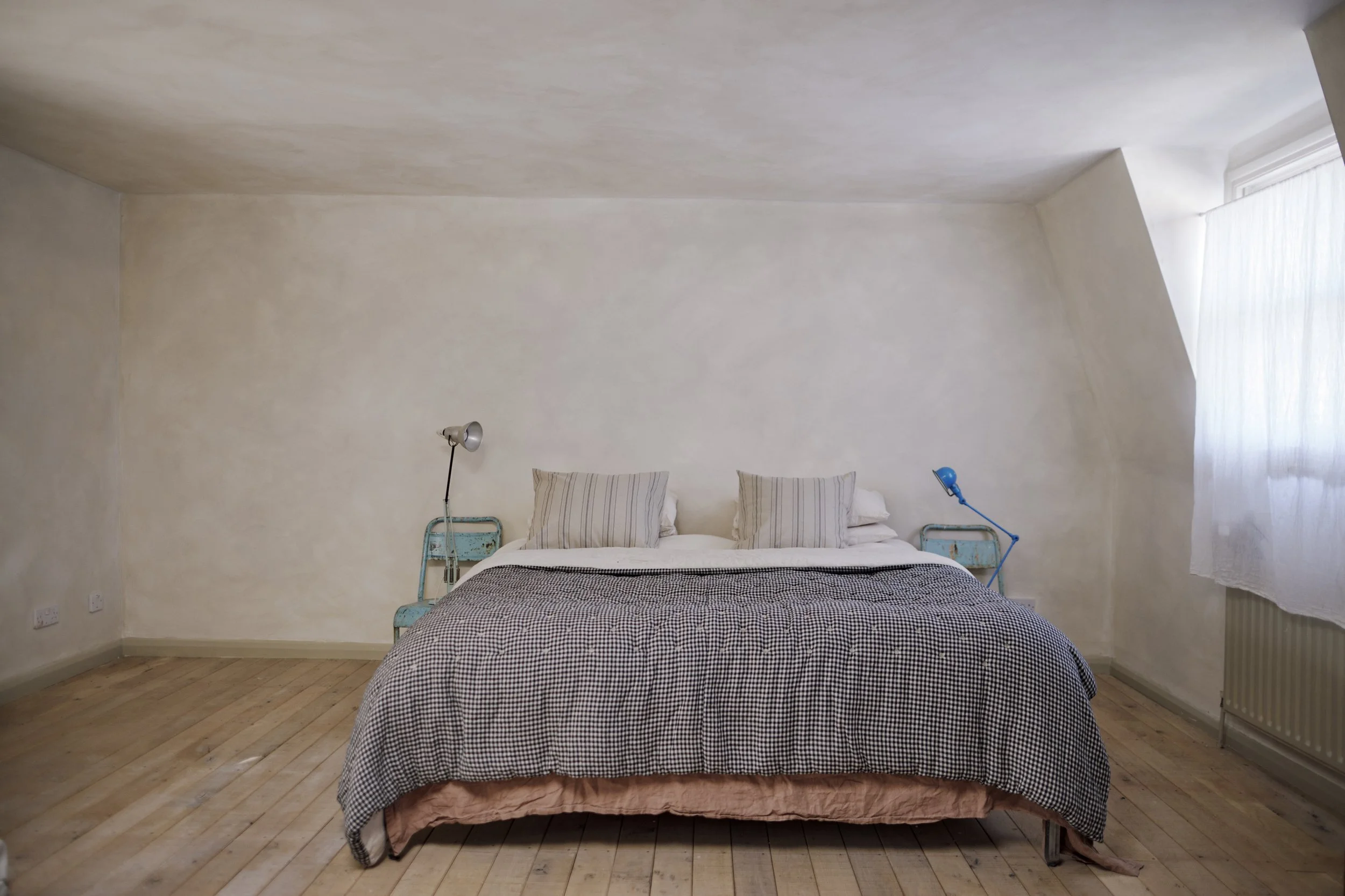 A bedroom with a bed featuring a checkered duvet cover, two striped pillows, and a pink bed skirt. Two vintage blue bedside tables with adjustable reading lamps are on either side of the bed. A window with sheer white curtains lets in natural light t