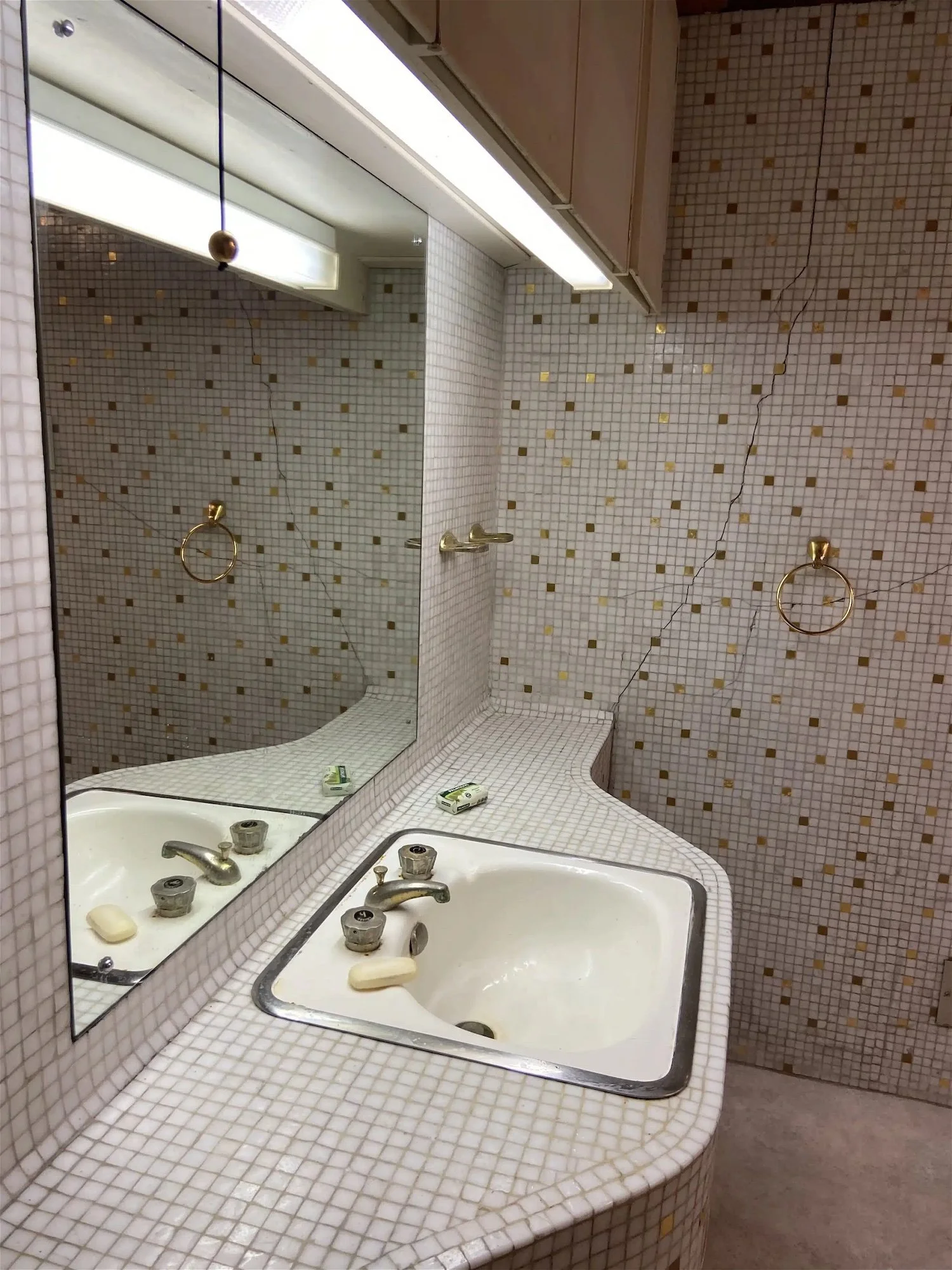 Restroom with a square sink, soap, and a large mirror. The walls are covered with small square mosaic tiles in white, gold, and brown. There are gold towel rings on the wall, and a cabinet mounted above the sink. The countertop is tiled with small wh