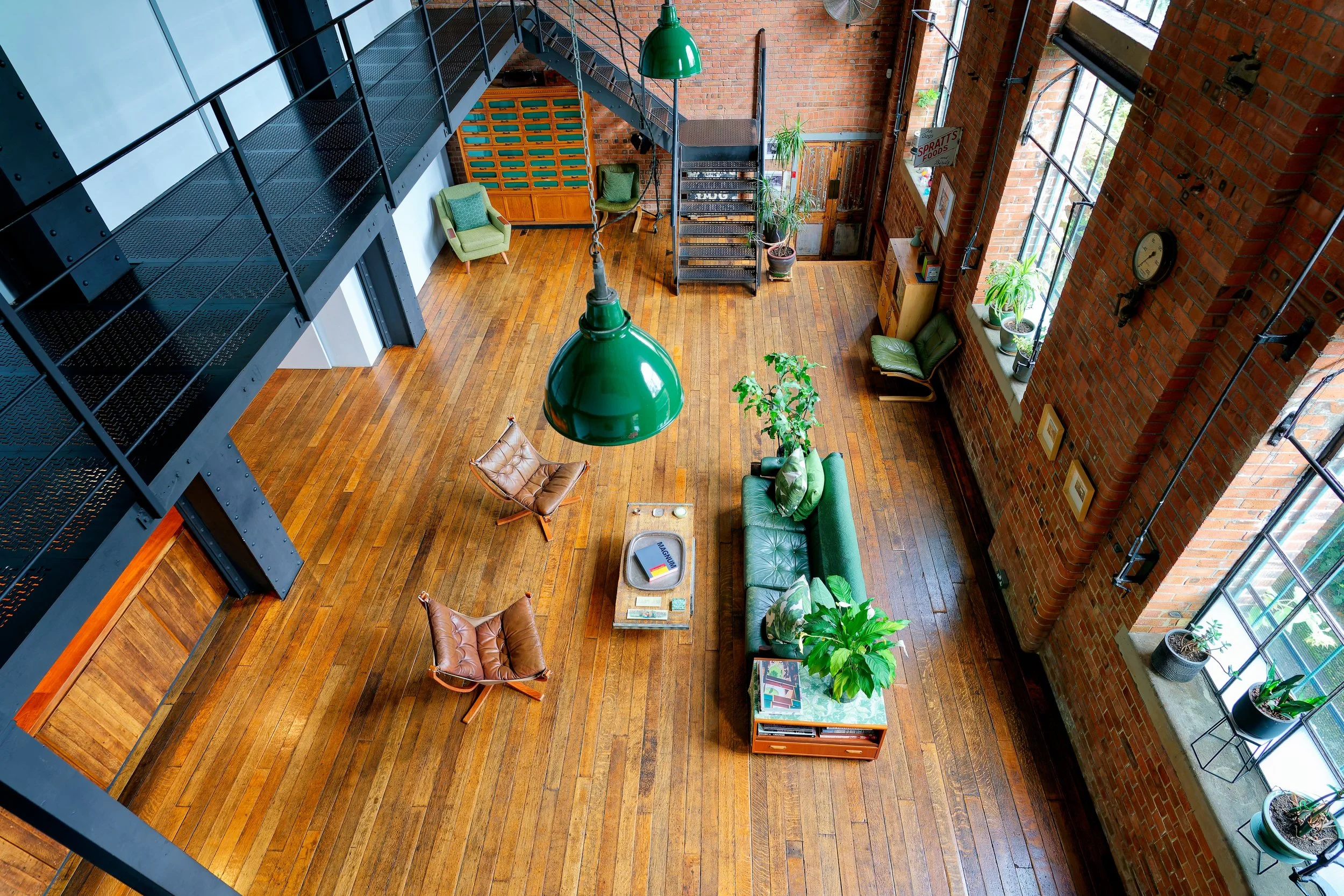 A spacious industrial-style loft living room with wooden flooring, large windows with potted plants, a green leather sofa, brown leather chairs, a coffee table, and a staircase leading to a mezzanine level.