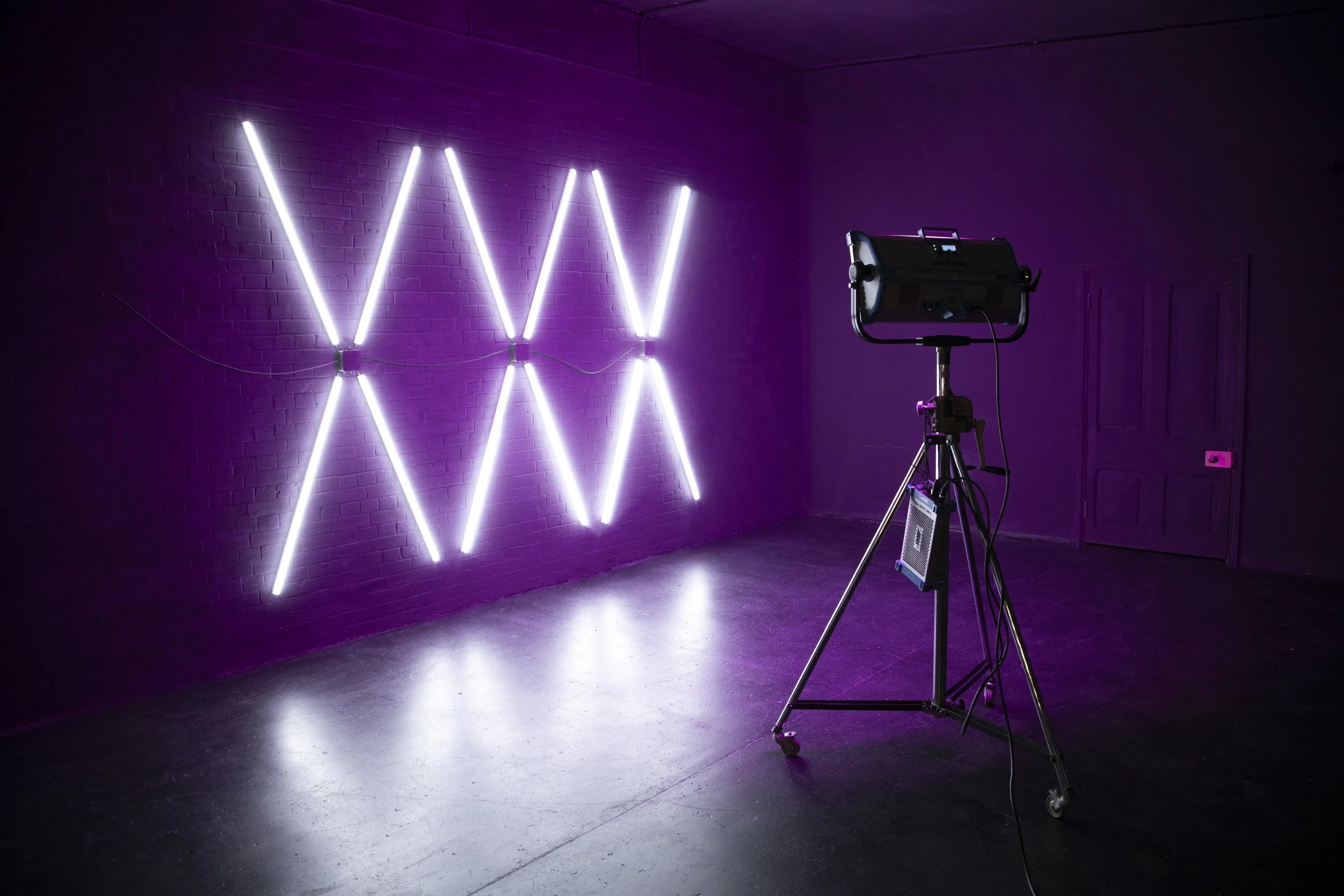 A room with a purple brick wall illuminated by a purple neon light installation spelling out the word "WOW" with vertical and diagonal neon tubes. In front of the wall, there is a photography or videography lighting setup on a tripod.