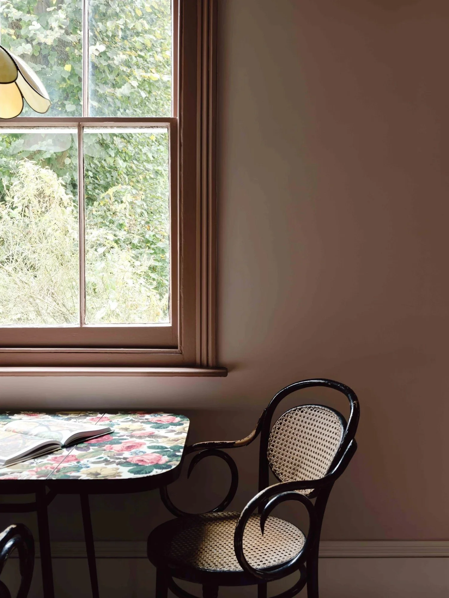A window with wooden trim letting in natural light, a floral-patterned table with an open book, and a black wicker chair with a cane backrest on a beige wall.