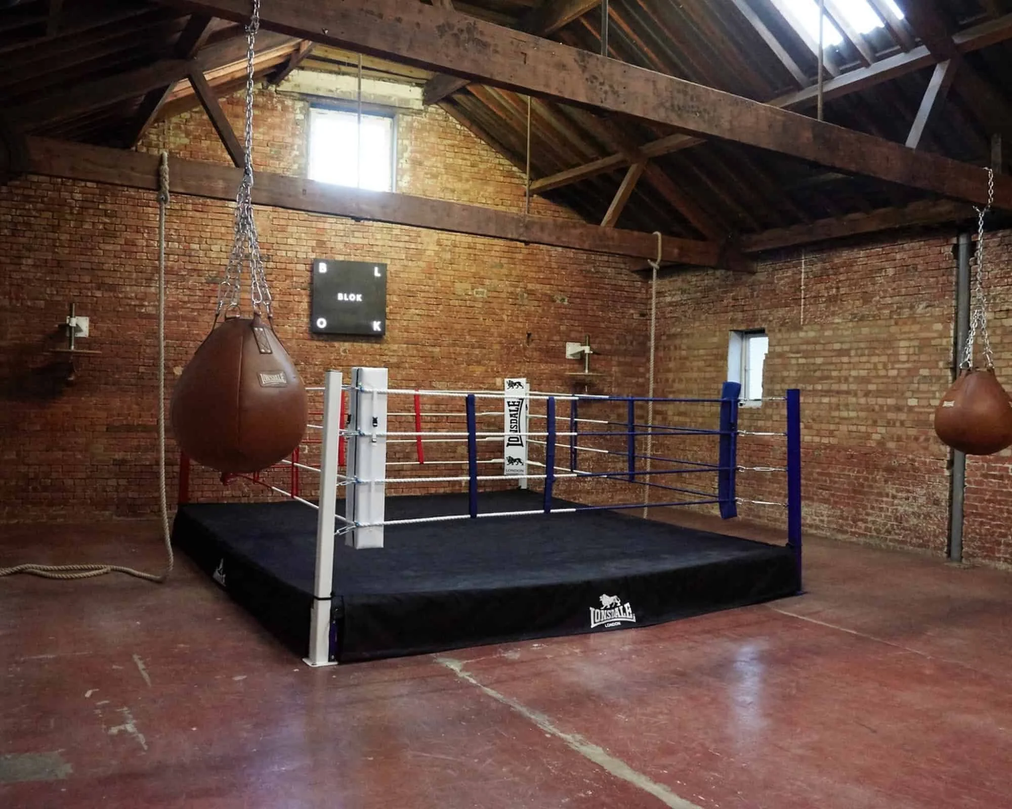 An old boxing gym with brick walls, a boxing ring in the center, and two punching bags hanging from the ceiling, with exposed wooden beams.