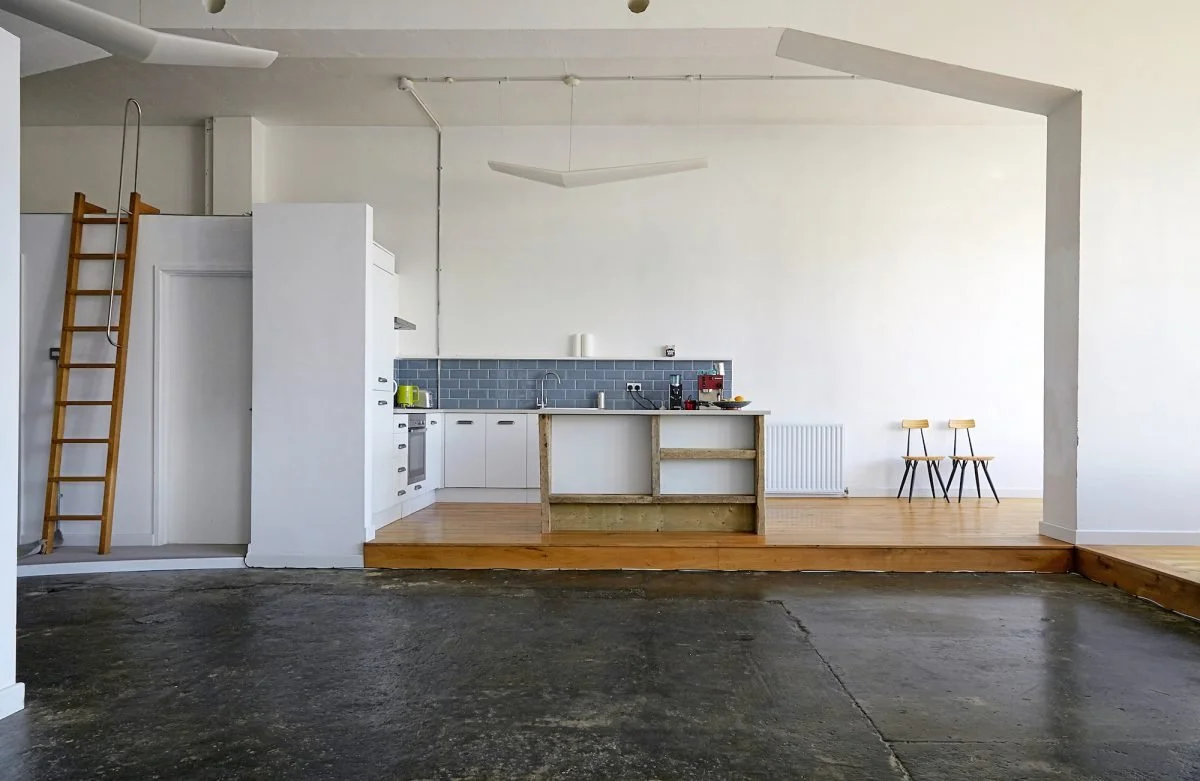 Open-concept kitchen with white cabinets, gray tile backsplash, and a small breakfast bar under construction. To the right, a small dining area with two chairs. The floor shifts from concrete in the foreground to wood in the kitchen and dining area.