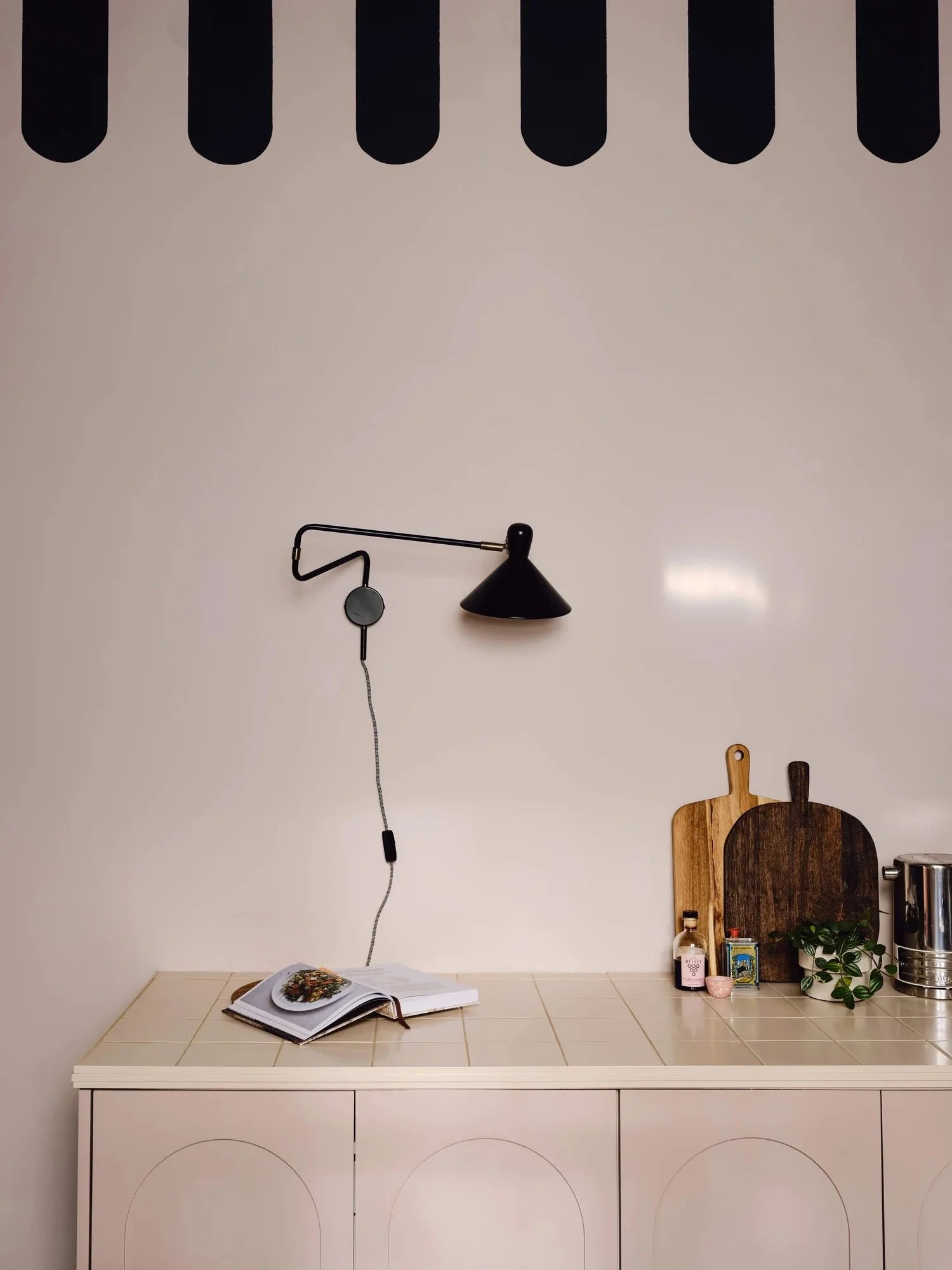 Kitchen countertop with open cookbook, wooden cutting boards, a small plant, spice jar, and a metal appliance, with a black wall-mounted lamp and a beige wall in the background.