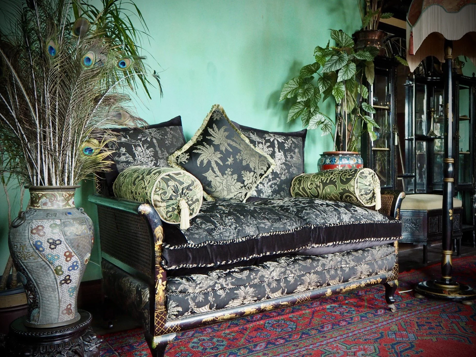 A vintage black and gold sofa with floral patterns, topped with green and black pillows, flanked by large potted peacock feather and leafy plants, set against a mint green wall, with a glass display cabinet and a colorful rug completing the cozy inte