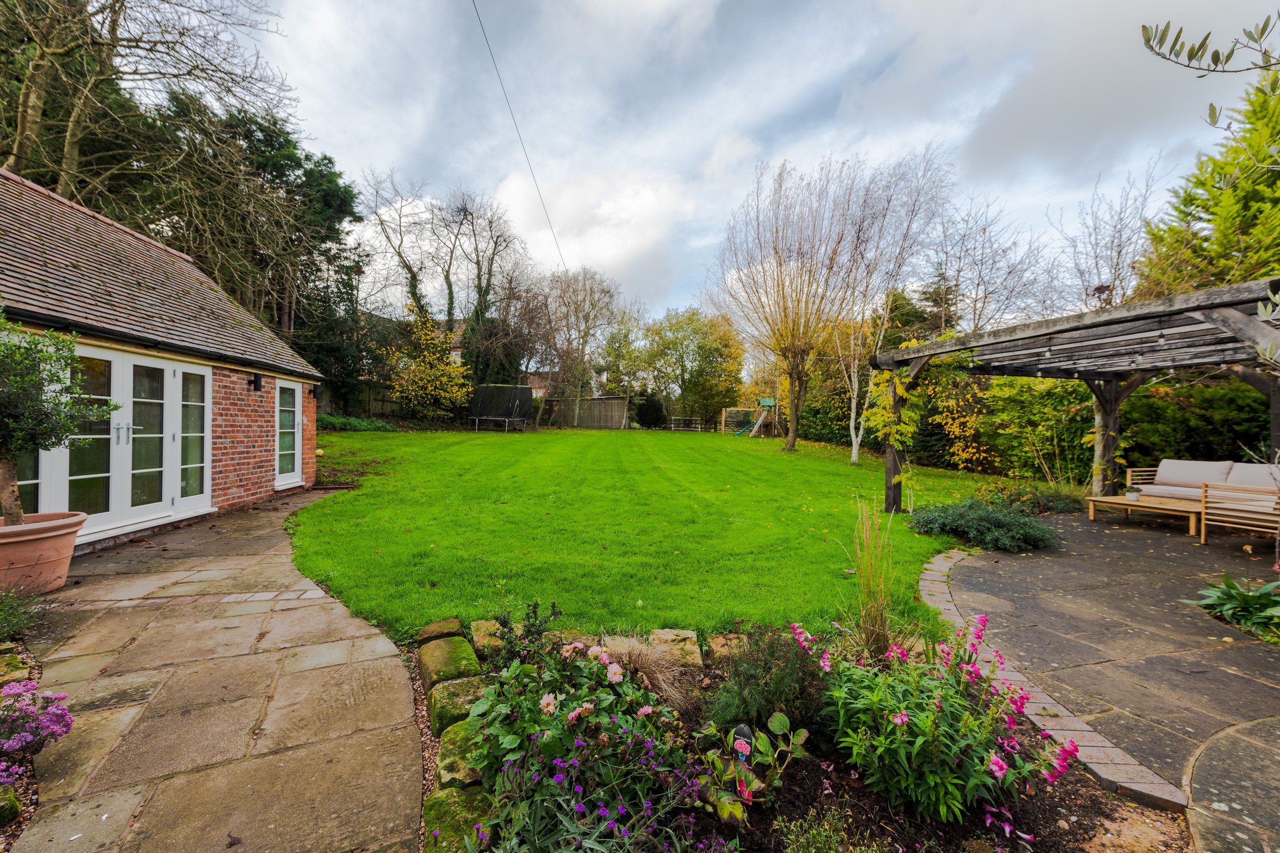 A backyard garden with a brick house to the left, a green lawn, and various trees and plants, including pink and purple flowers in the foreground. There is a paved stone patio with a wooden bench and a pergola on the right.
