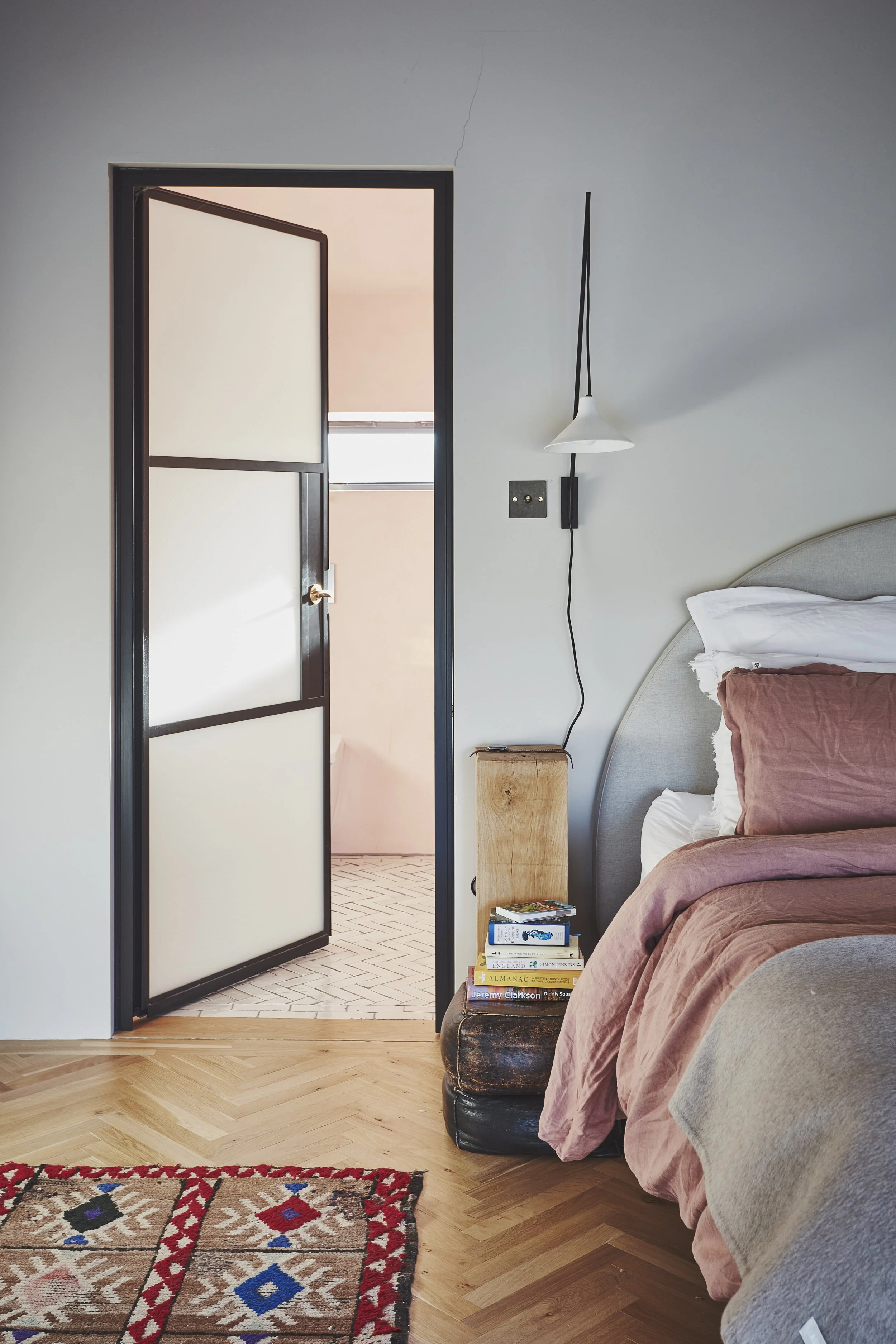 A bedroom with a bed covered in pink and beige bedding, a black nightstand with books, a small wooden block, a wall-mounted lamp, a door with frosted glass panels, and a patterned rug on a wood floor.