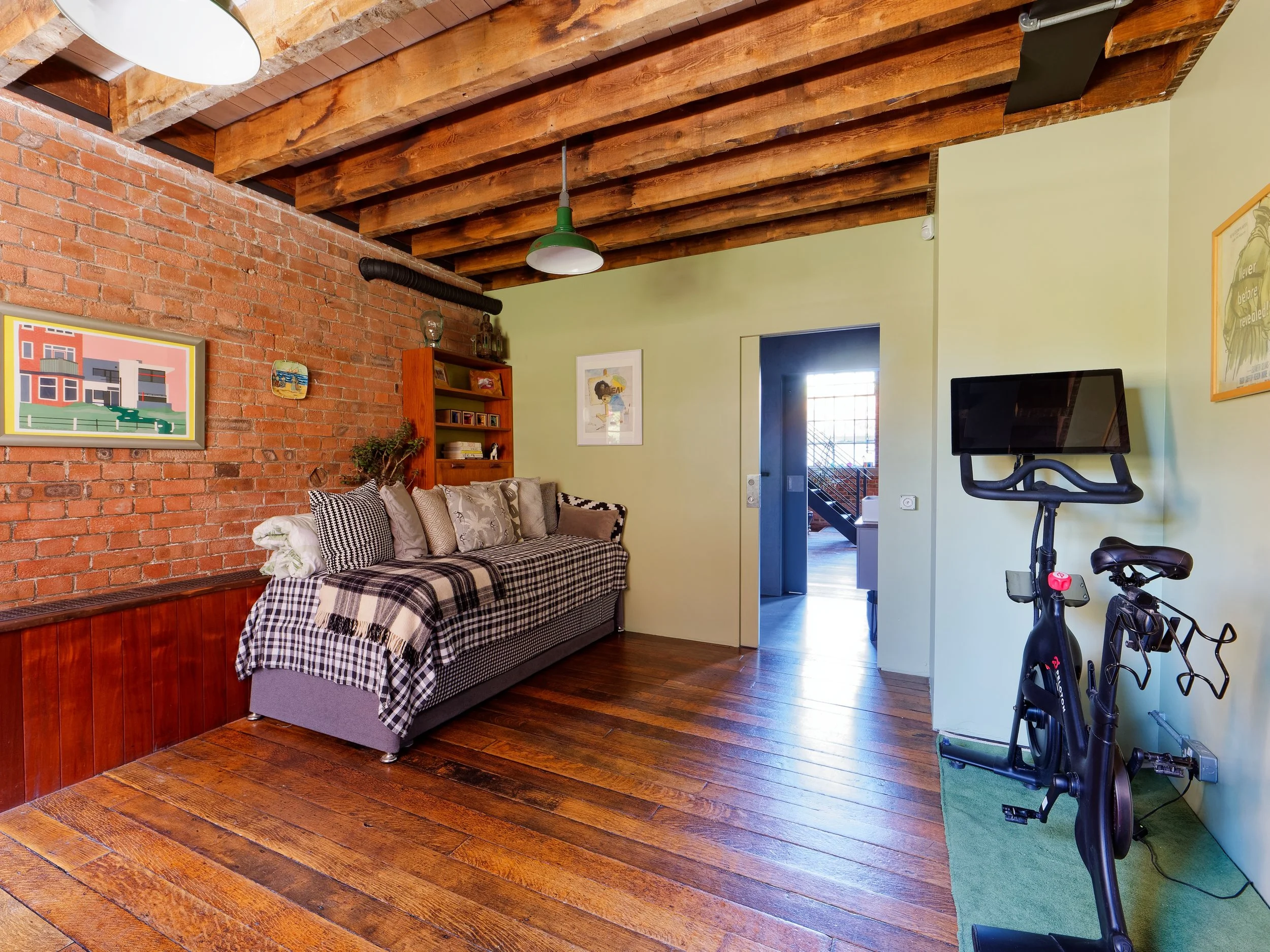Living room with wooden floors, brick wall, green painted walls, and exposed wooden beams on the ceiling. Contains a checkered sofa with pillows, a wooden shelf, framed artwork on the walls, a stationary exercise bike, and an entrance to another room