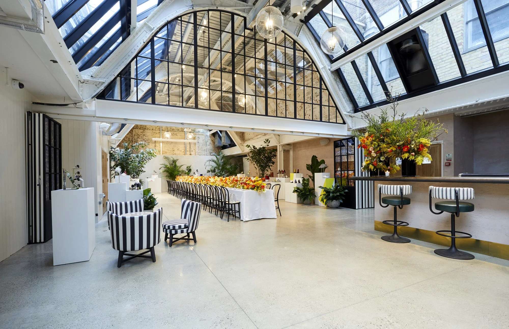 Interior of a bright, modern event space with a glass ceiling, decorated with flowers and plants, featuring a long table with floral arrangements and seating for a celebration.
