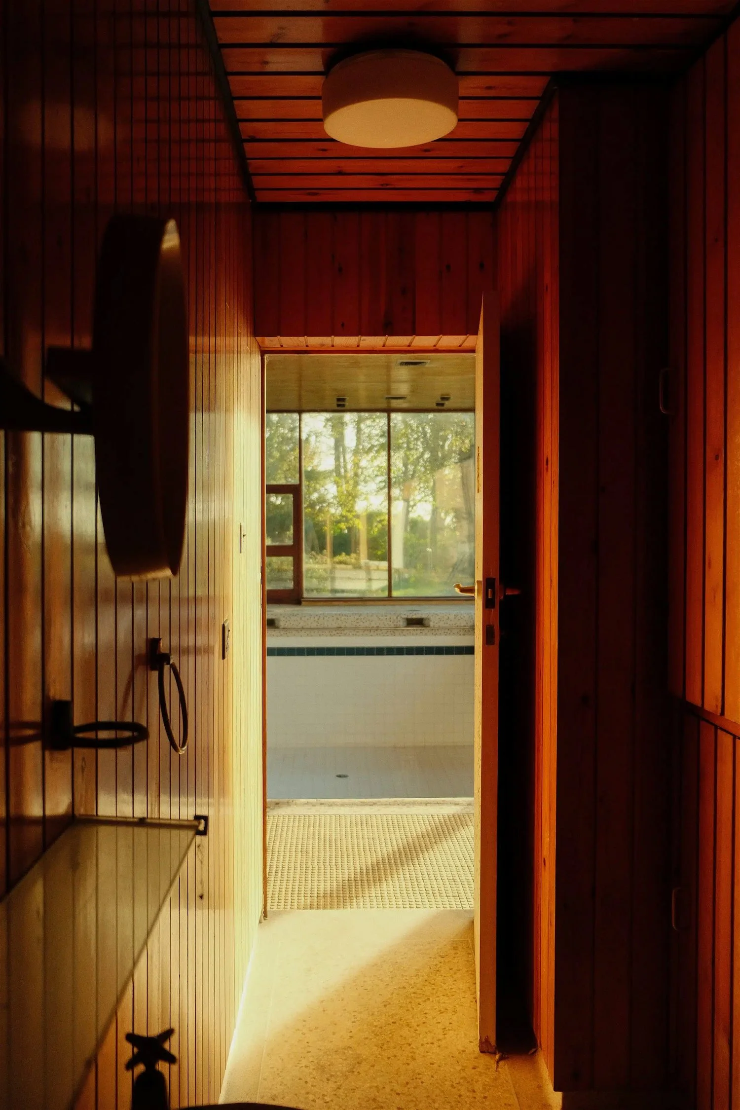 View through a doorway into a sunlit indoor swimming pool area with large windows and green trees outside.