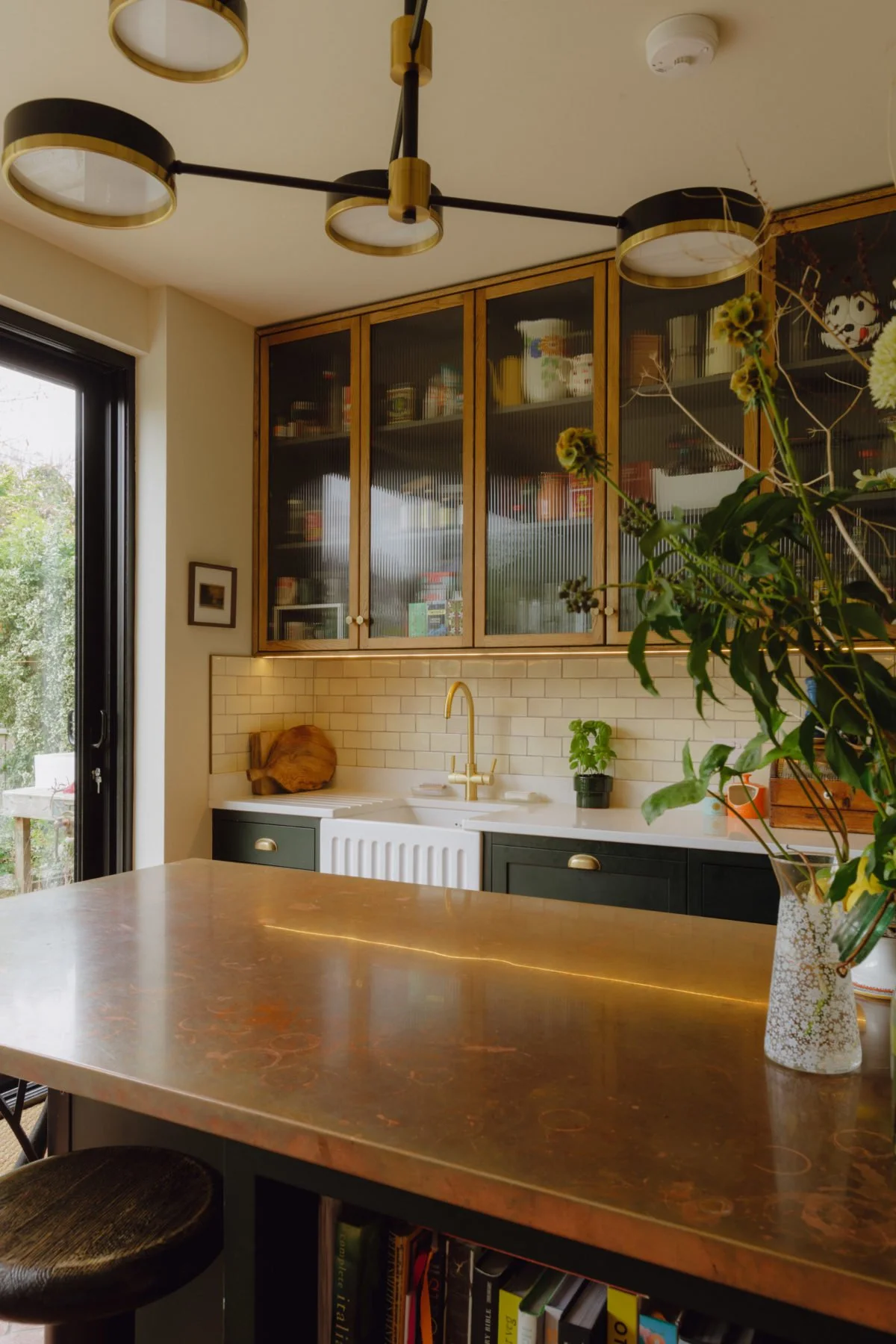A modern kitchen with dark green cabinets, a white subway tile backsplash, a brass faucet, a wooden cutting board, and a large window with a view of trees. A vase with flowers on the counter and a black and gold ceiling light fixture.