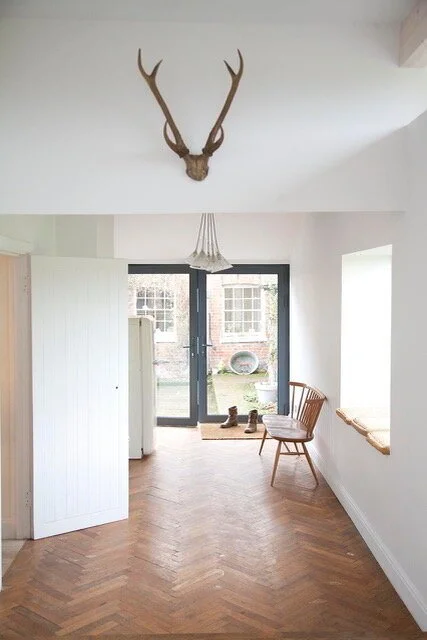 Interior of a room with a deer antler mounted on the wall, a glass door with shoes outside, a wooden chair, and a window seat.