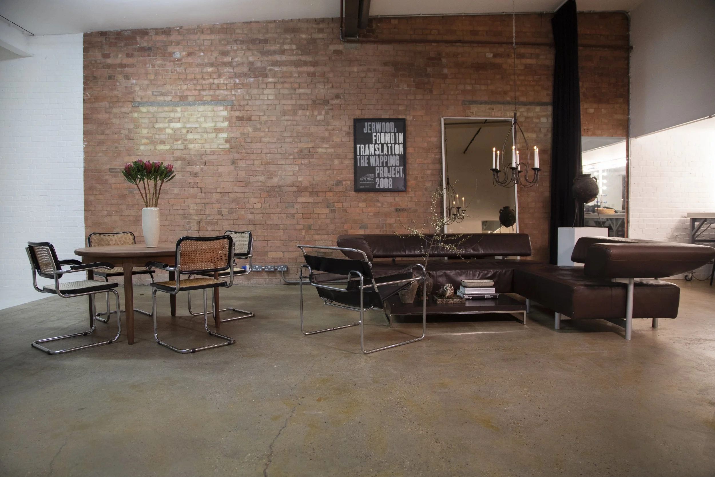 An industrial-style living room with exposed brick walls, a round wooden dining table with five chairs, a dark brown leather sofa with a matching chaise lounge, a large mirror on the wall, a chandelier with candles, and a vase of flowers on the table