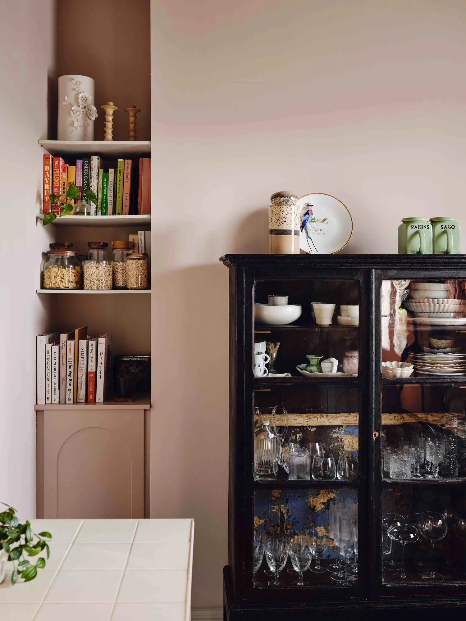 A kitchen corner with a white shelf filled with cookbooks, jars of dry food, and decorative items next to a black display cabinet with dishes, glasses, and bowls. There are decorative jars on top of the cabinet and a plate with a bird illustration.