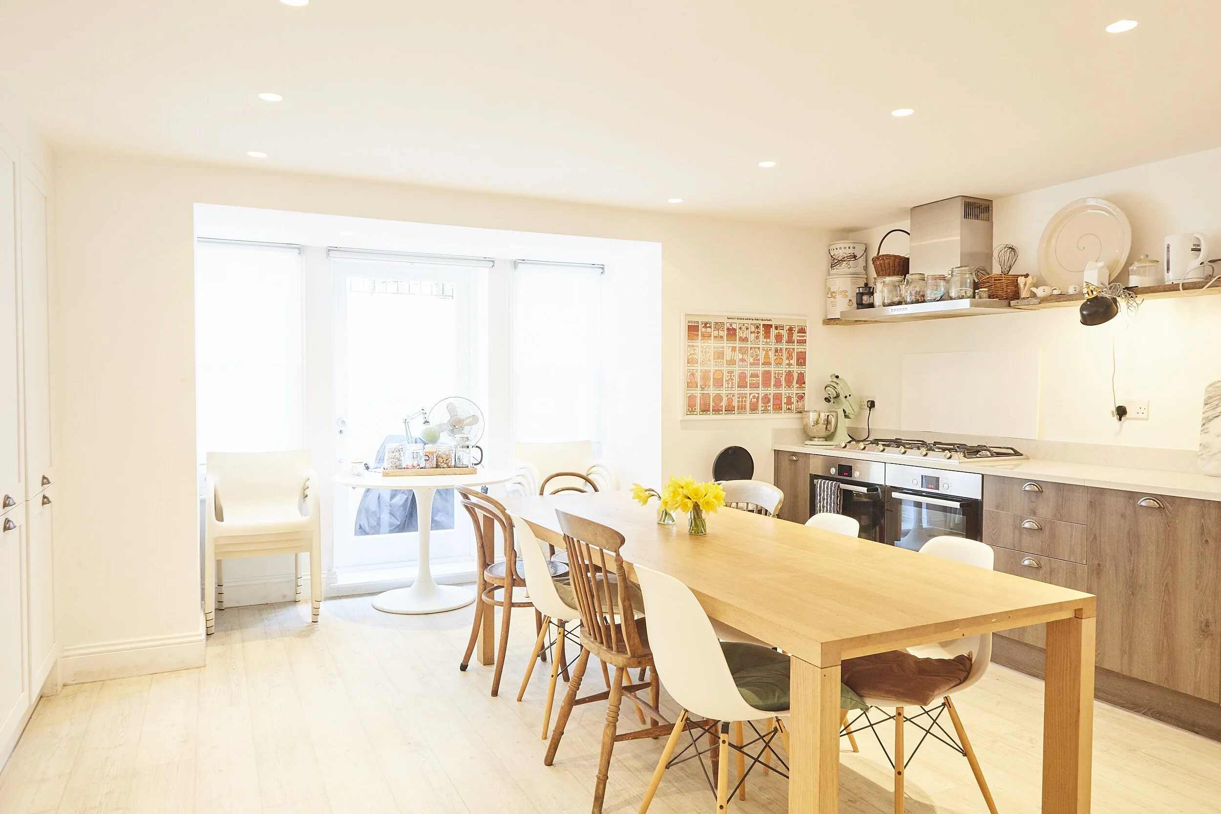 Bright, modern kitchen and dining area with a wooden table, various chairs, a bouquet of yellow flowers, and kitchen appliances and shelves on the back wall.