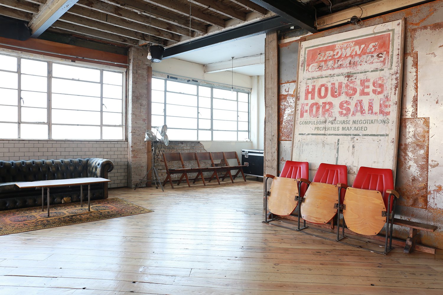 An empty industrial-style room with hardwood floors, large windows, a black leather couch, red chairs folded and stacked, a vintage sign reading 'Houses for Sale,' and various furniture and decor elements.