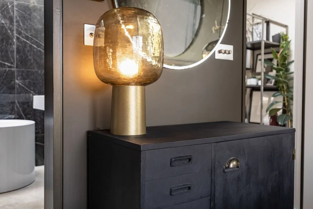 A modern interior decor scene featuring a black wooden sideboard with a brass and glass table lamp on top, a wall-mounted round mirror with LED lighting, and a plant in the background.