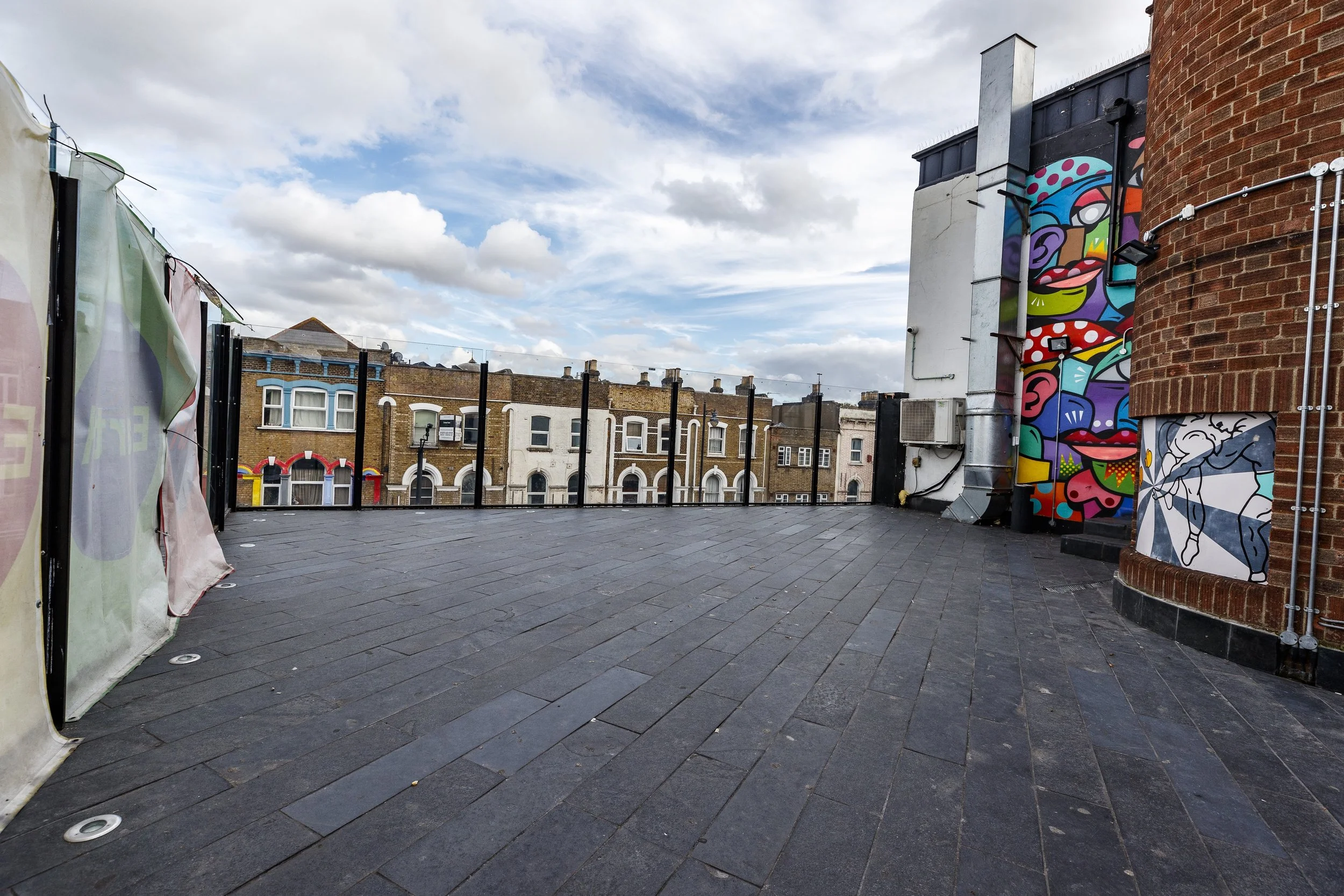 Rooftop terrace with black tiled flooring, colorful murals on the walls, and a view of neighboring brick buildings under a partly cloudy sky.