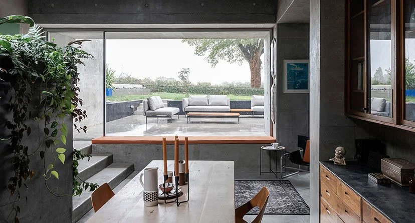 Modern dining room with a view of an outdoor patio and garden, featuring a white dining table, wooden chairs, and decorative candles, with large windows and a concrete wall.