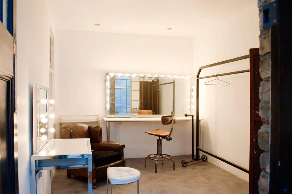 A backstage dressing room with illuminated vanity mirror, brown leather armchair, white table, red chair, freestanding clothing rack, large windowed mirror, and plain white walls.