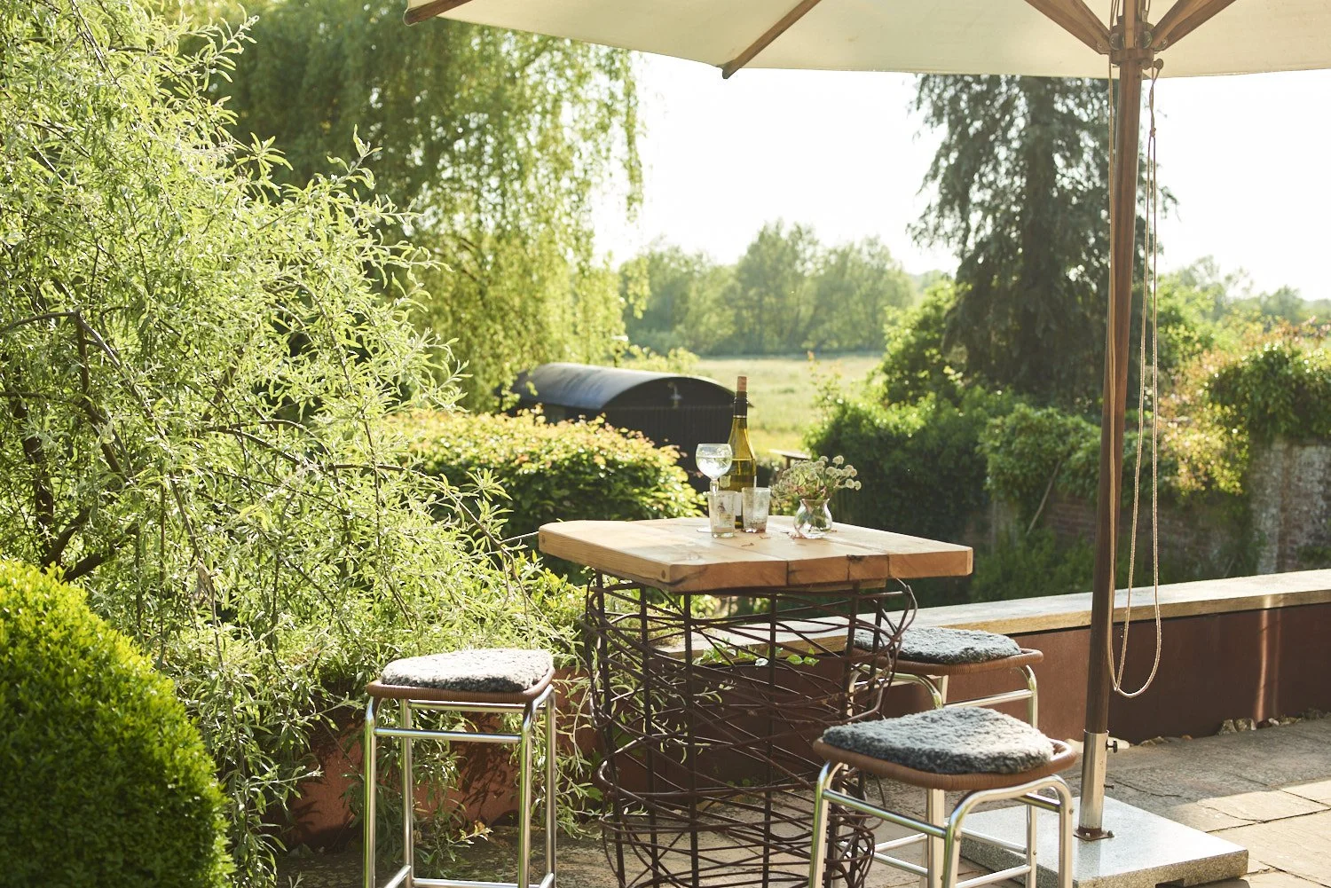 Outdoor patio table with chairs, glass of water, wine bottle, flowers, umbrella, lush green bushes, and trees in the background on a sunny day.