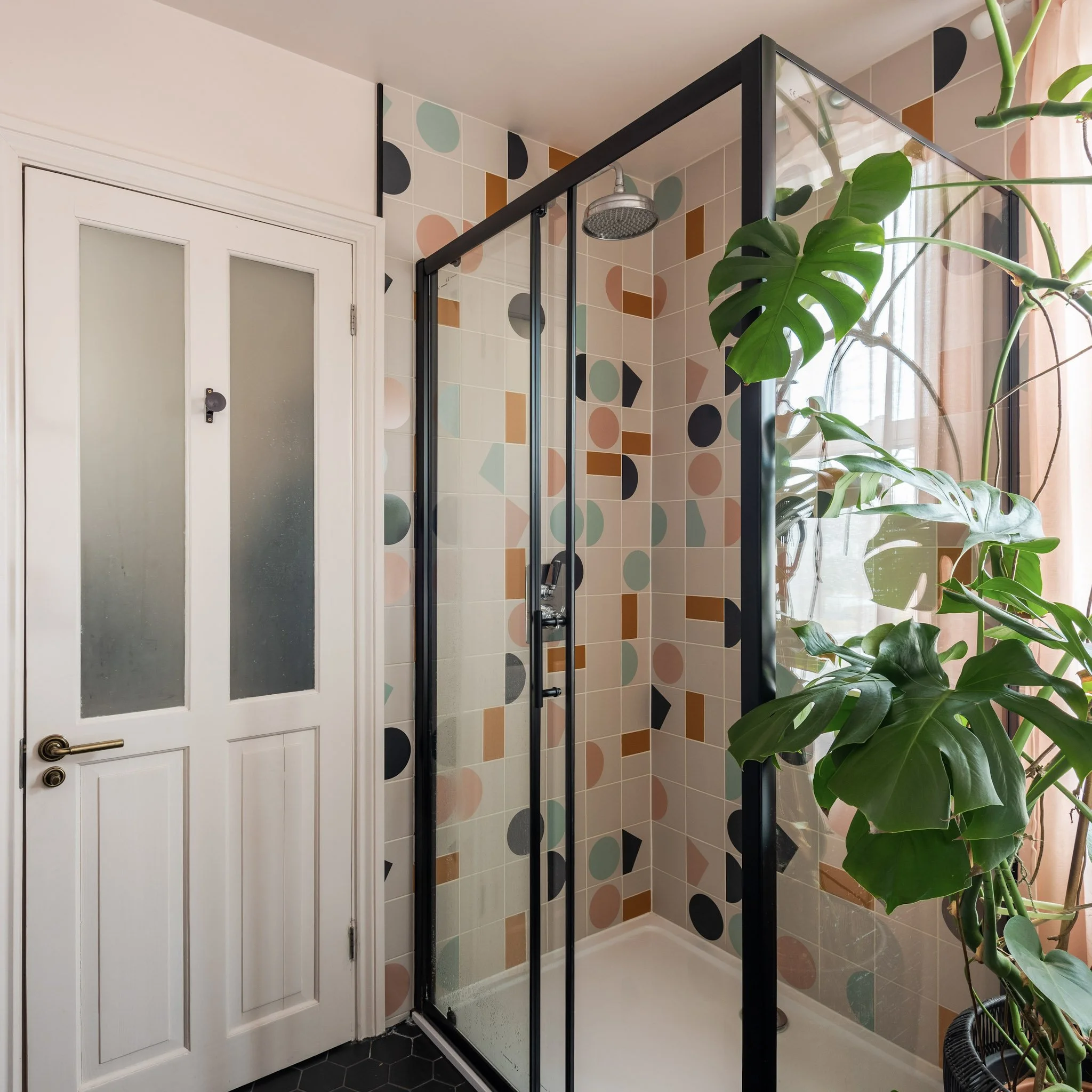 Small bathroom shower area with colorful geometric tile pattern, featuring a black-framed glass shower enclosure, a rain shower head, a white door with frosted glass panels, and large green indoor plants beside the window with sheer curtains.
