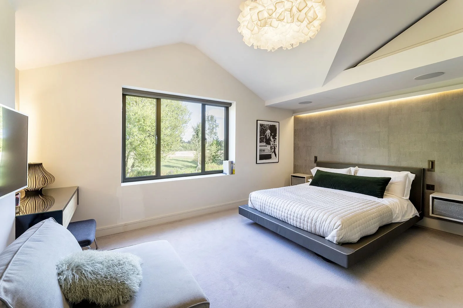 Modern bedroom with large bed, white bedding, green accent pillow, beige carpet, window with green trees outside, black and white wall art, beige textured wall, and contemporary lighting fixtures.