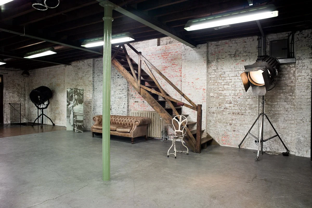 Empty photography studio with brick walls, large studio lights, vintage sofa, white chair, and wooden staircase.
