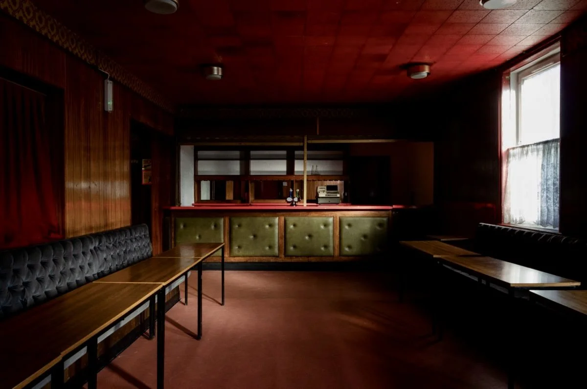Empty vintage bar with wooden paneling, red ceiling, dark tables, tufted booths, and a window with curtains