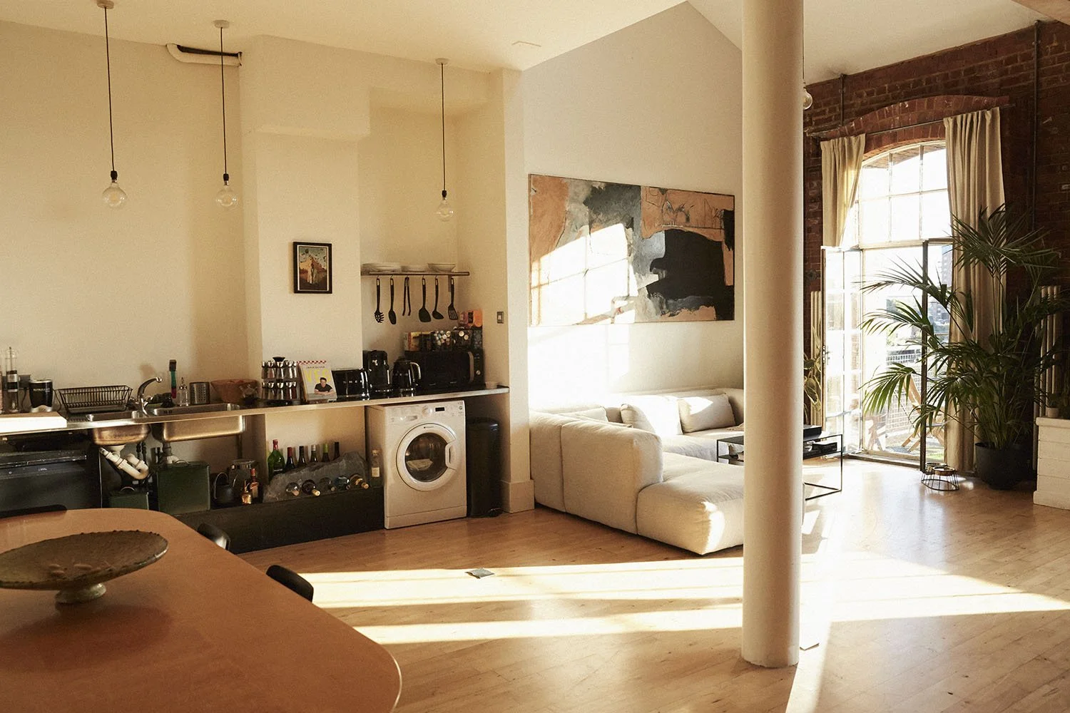 Bright living room with white sectional sofa, large window with curtains, wooden flooring, and exposed brick wall, with plants and artwork, and sunlight streaming in.