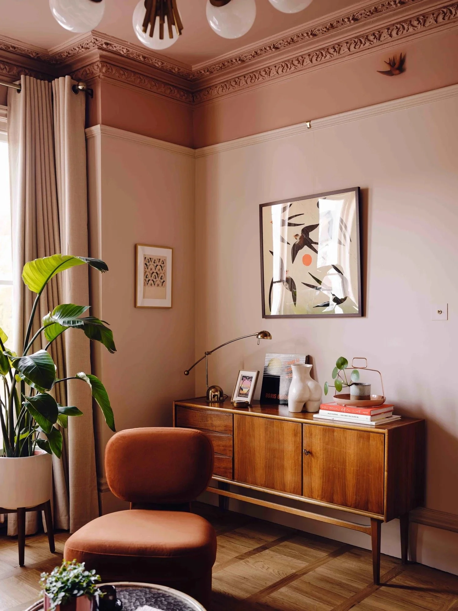 A cozy living room corner with a vintage wooden sideboard, a peach-colored velvet armchair, and a large potted plant near a window with beige curtains. Decor includes framed art, a white vase, books, and small decorative objects, with ornate crown mo