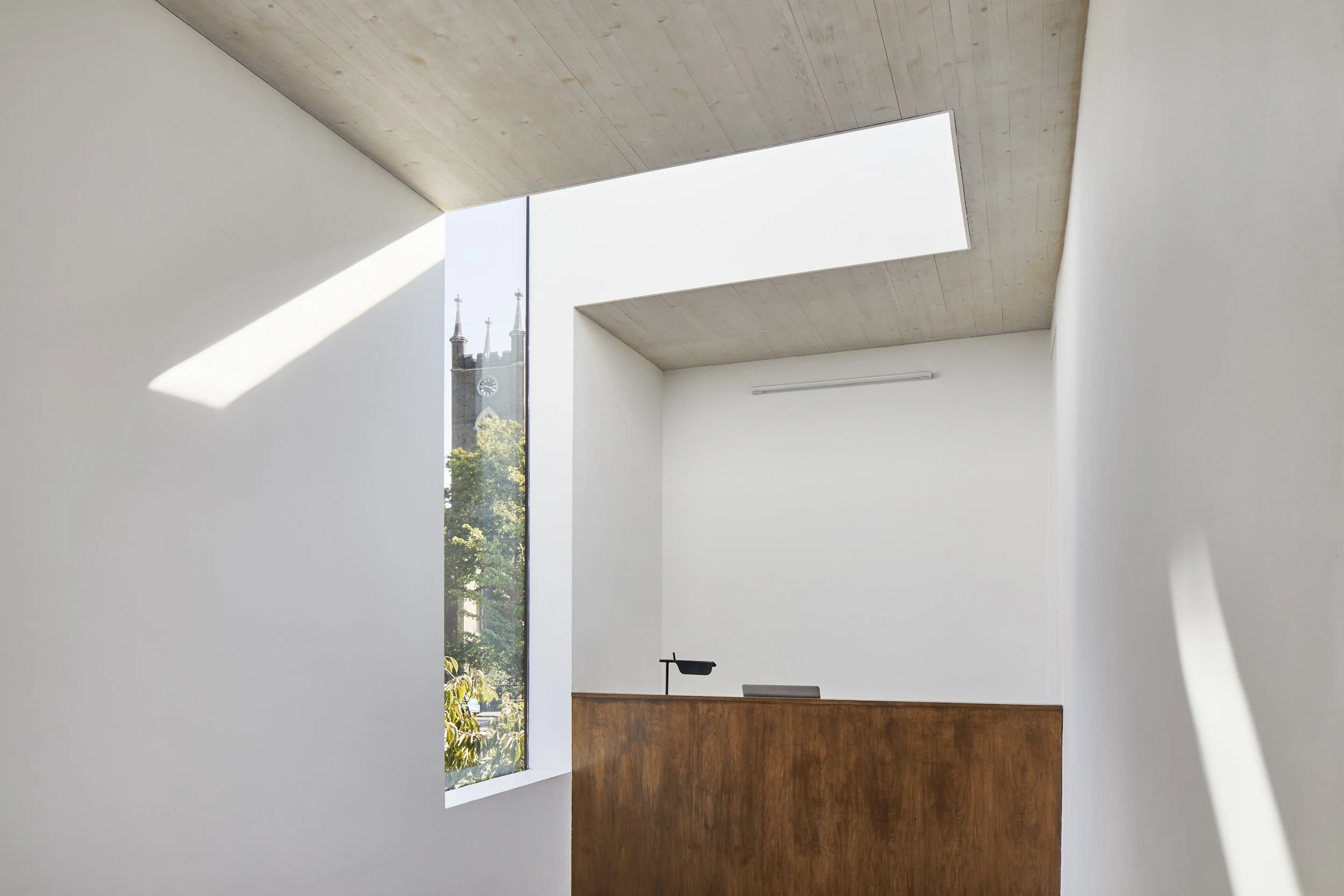 Minimalist interior space with white walls, wooden ceiling, large windows, and a wooden reception desk. A church steeple and clock are visible outside through the windows.
