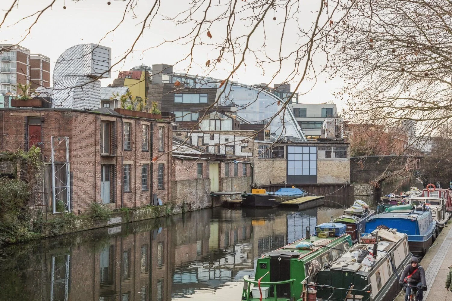 Boats docked along a canal with brick buildings and modern structures in the background, and leafless trees overhead.