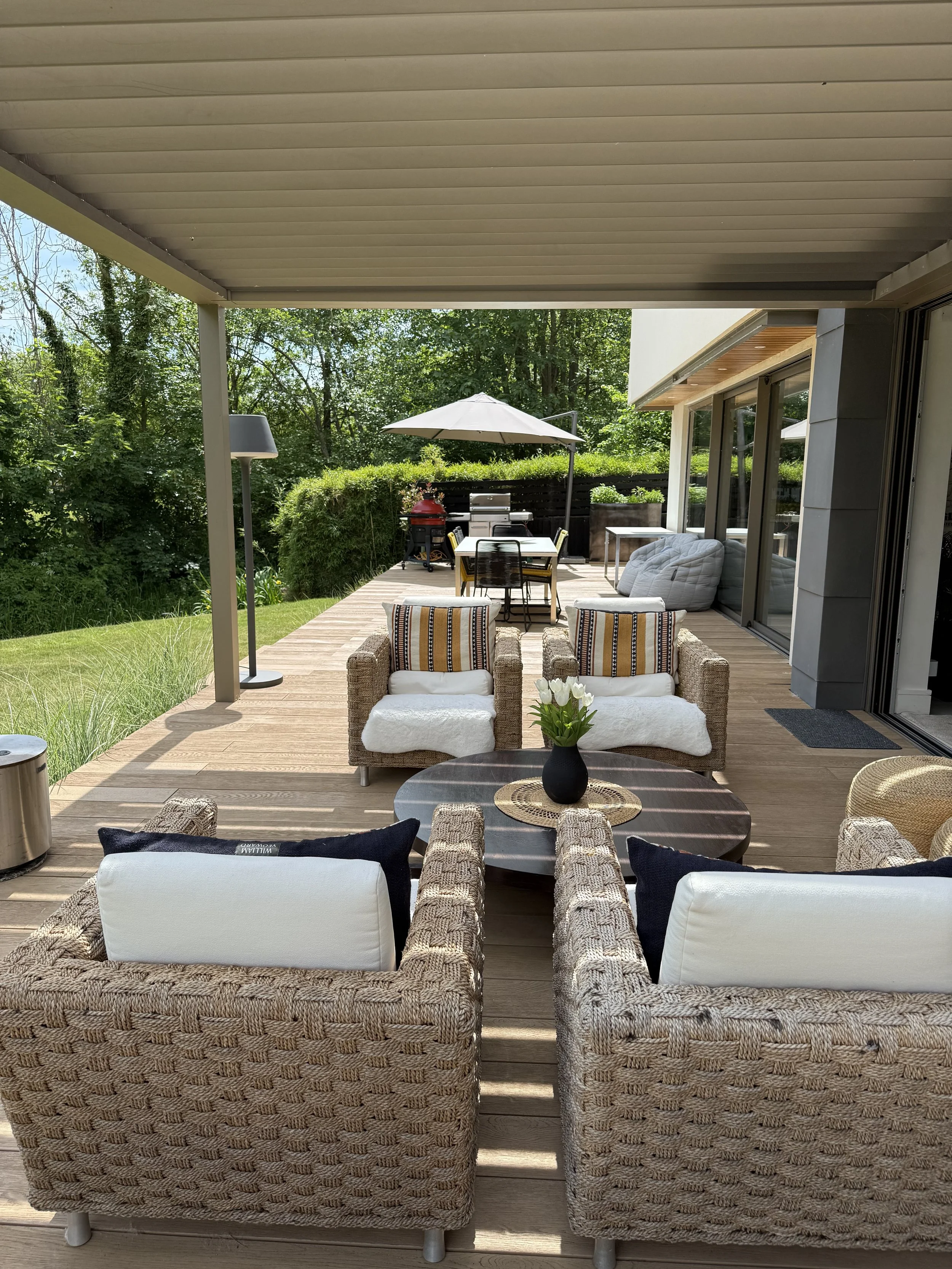 Outdoor patio with wicker and cushioned chairs, a round coffee table with a black vase of flowers, a dining table with yellow chairs and an umbrella, and lush green trees in the background.
