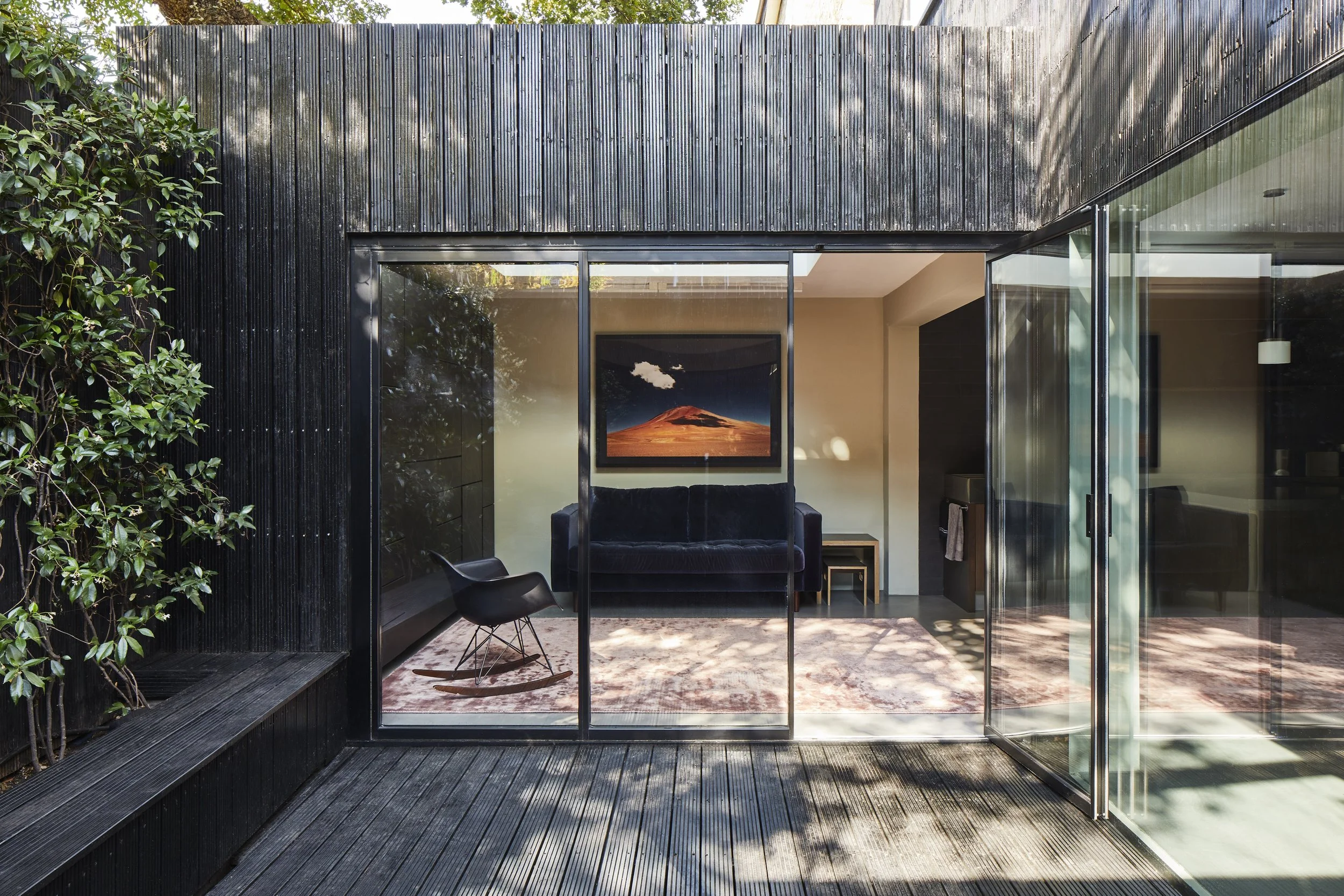 Modern house with a glass sliding door revealing a living room interior with a black sofa, a framed artwork of a mountain with a cloud, a chair, and a small side table inside. The exterior has a black wooden cladding and a wooden deck.