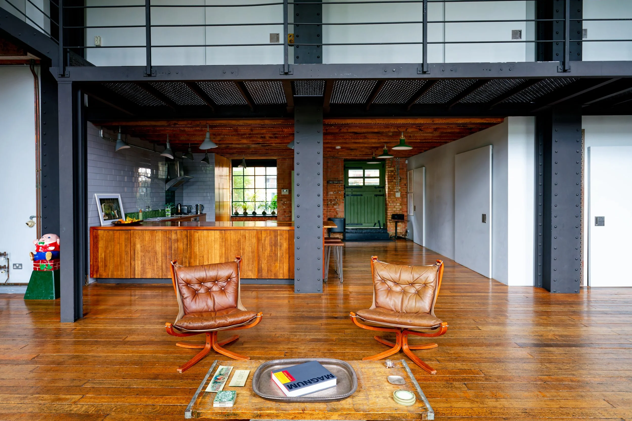 Open-plan living room with wooden floors, two leather lounge chairs, a coffee table with books and decorative items, an open kitchen with gray tiled walls, wooden cabinets, and a window with plants, brick and industrial design elements.