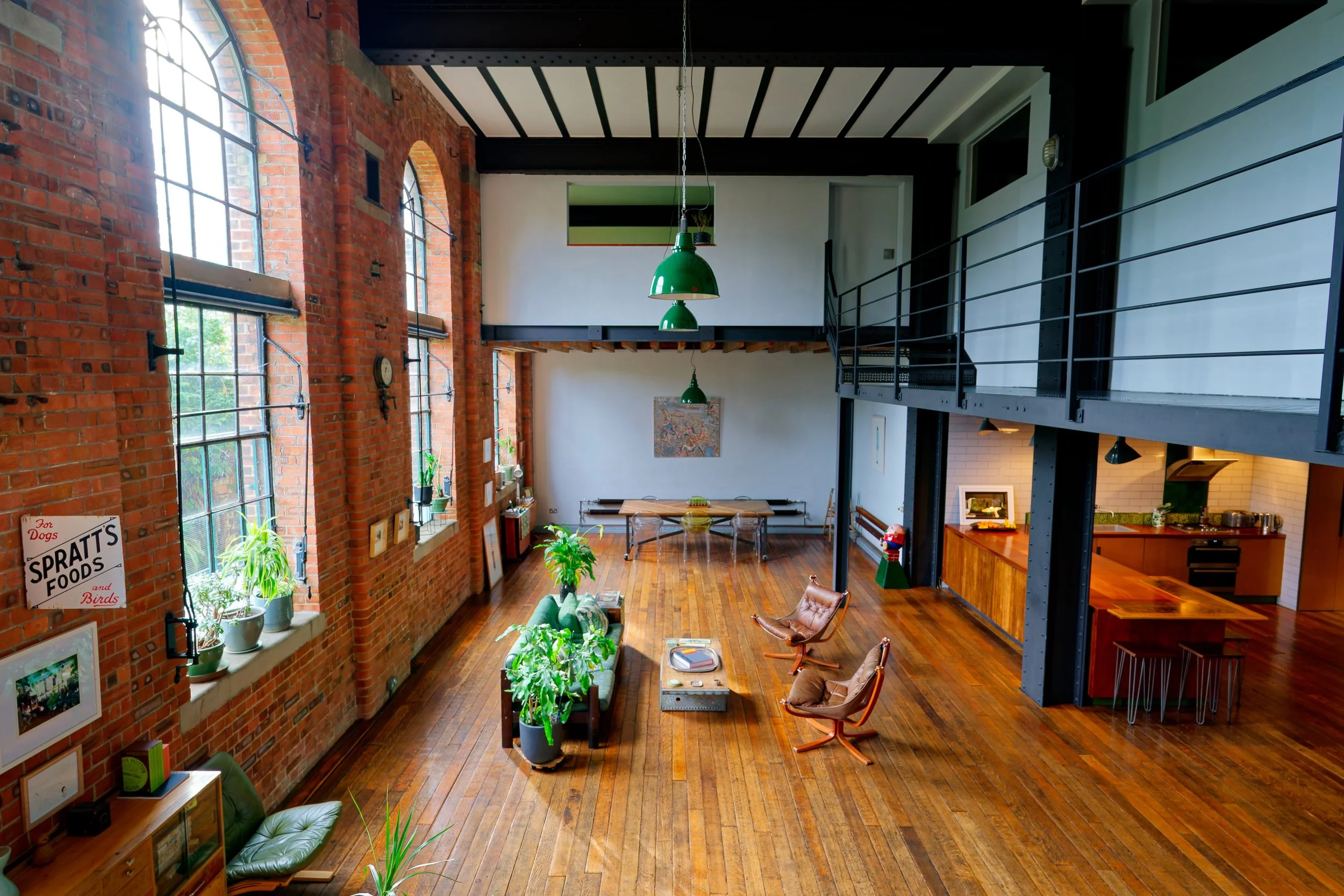 Spacious loft with exposed brick walls, tall windows, and hardwood floors, furnished with plants, a green lounge chair, two brown lounge chairs, a long dining table, and a kitchen area.
